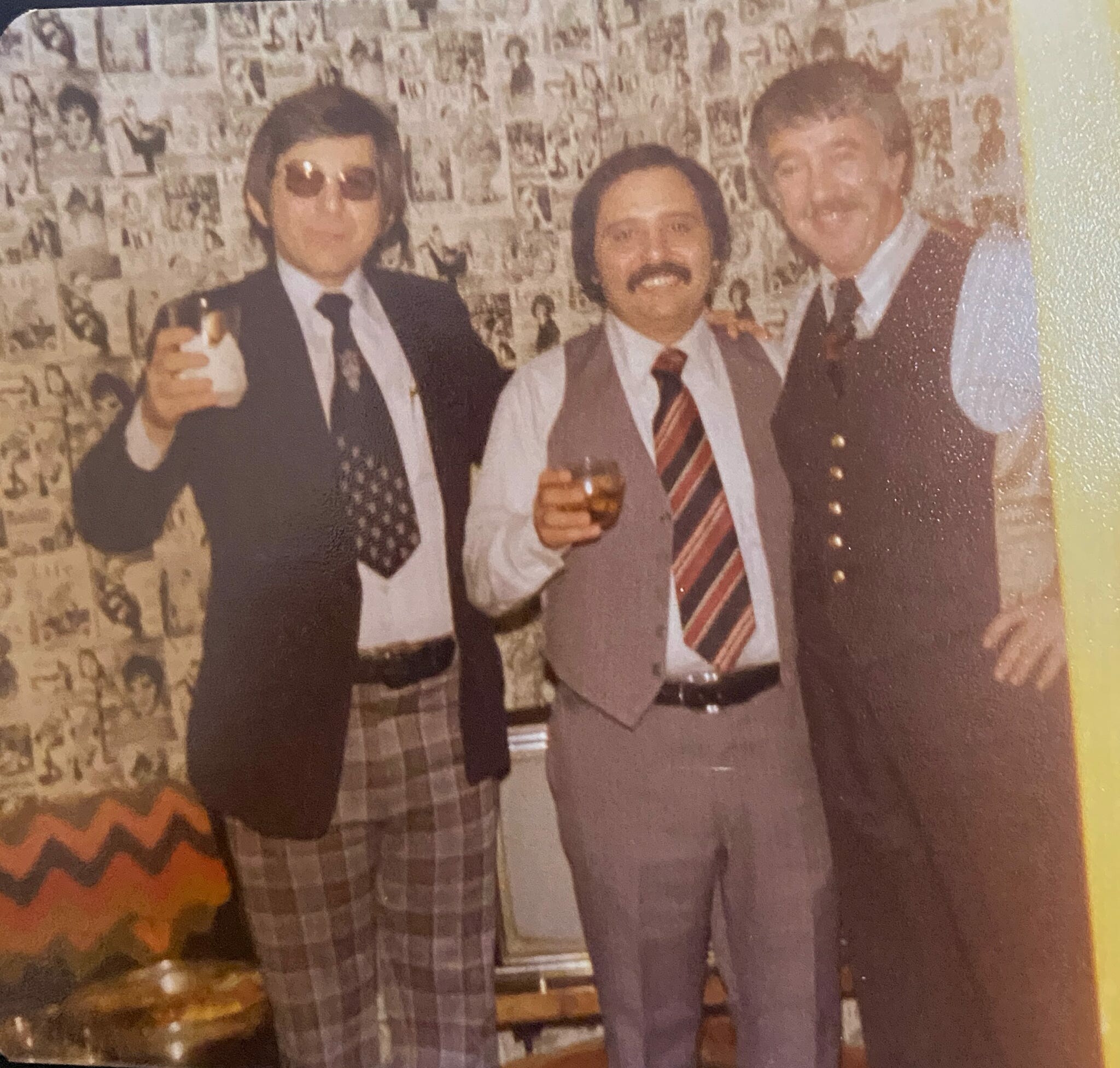 Three men in formal attire posing with drinks at a party, with a photo-covered wall in the background.