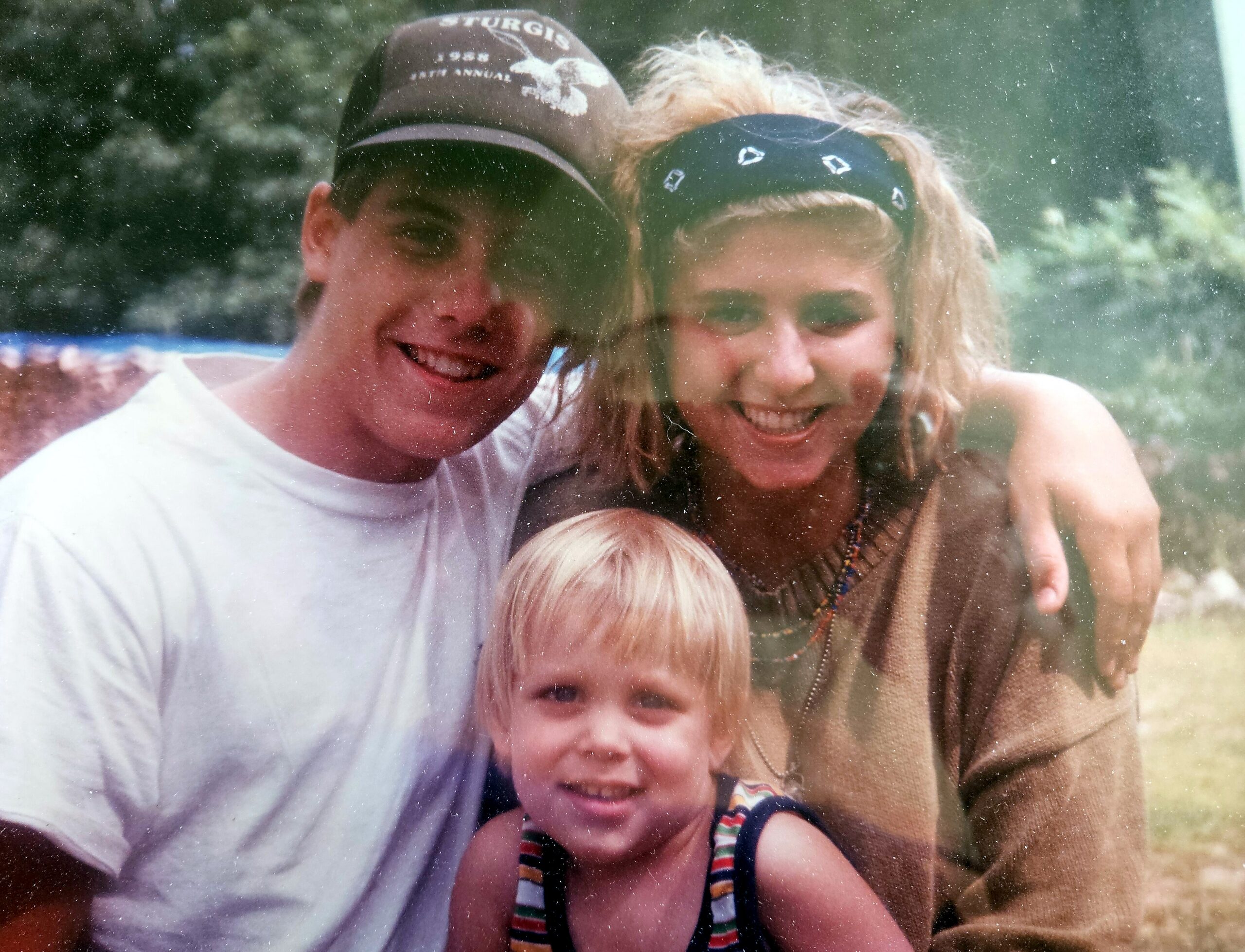 Three people outdoors, two adults and a child, smiling at the camera in a sunny setting.