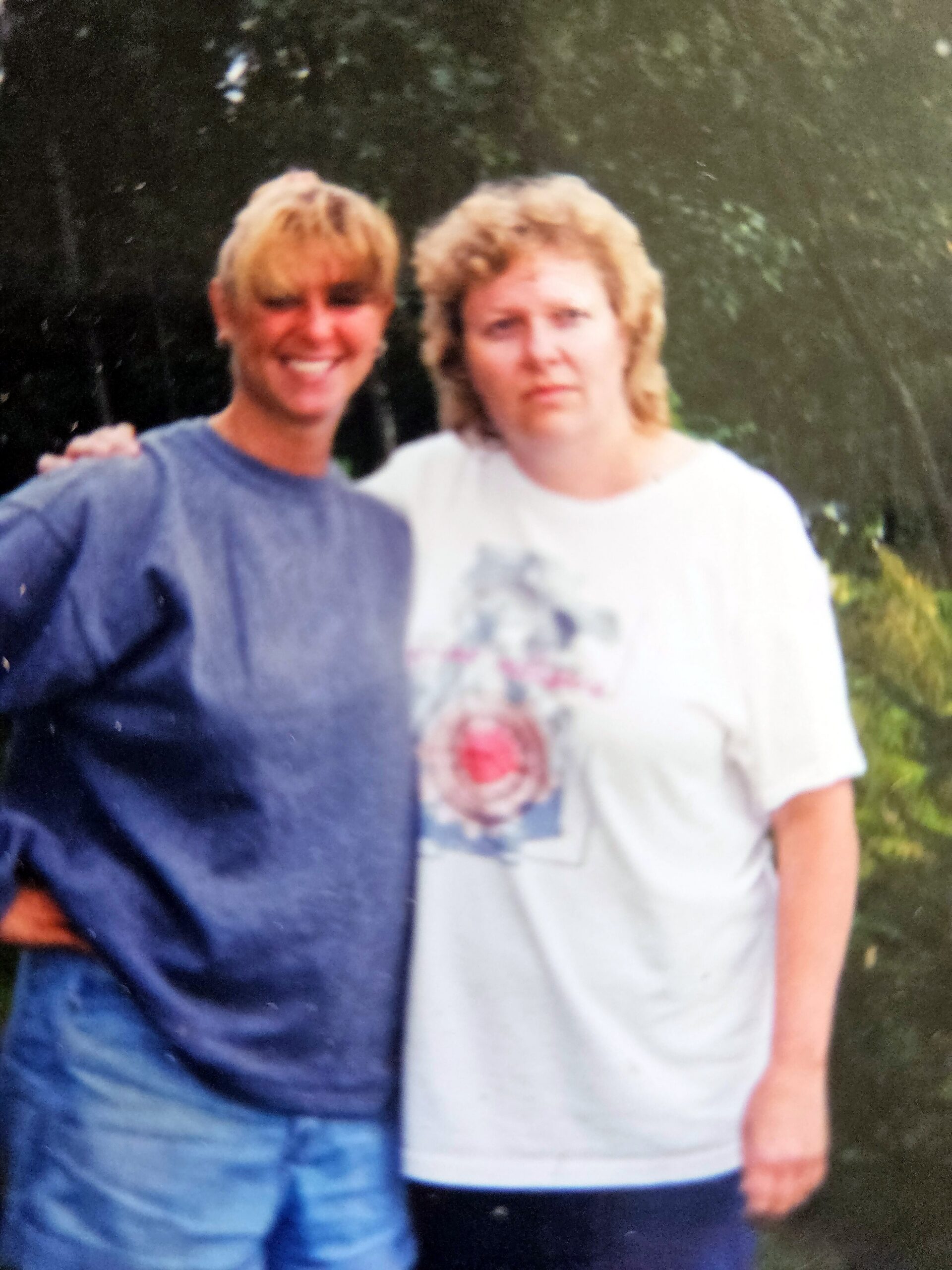 Two people posing together outdoors; left person in a blue shirt has an arm around the right person, who wears a light-colored T-shirt, both smiling.