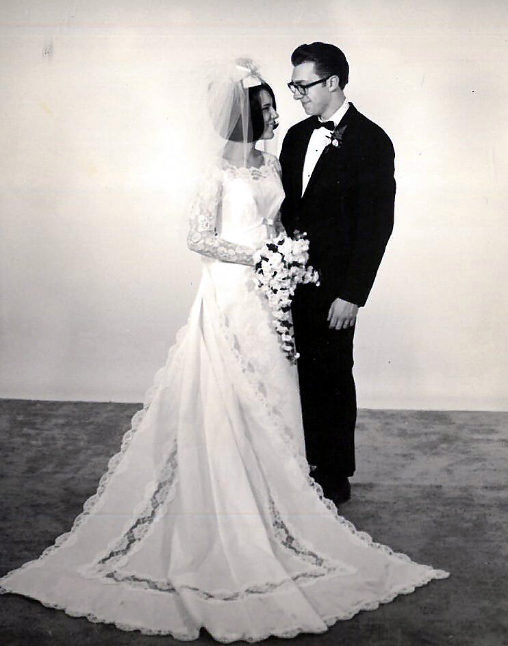 Bride wearing a long lace wedding gown and veil, holding a bouquet, standing with groom in a tuxedo and bow tie.
