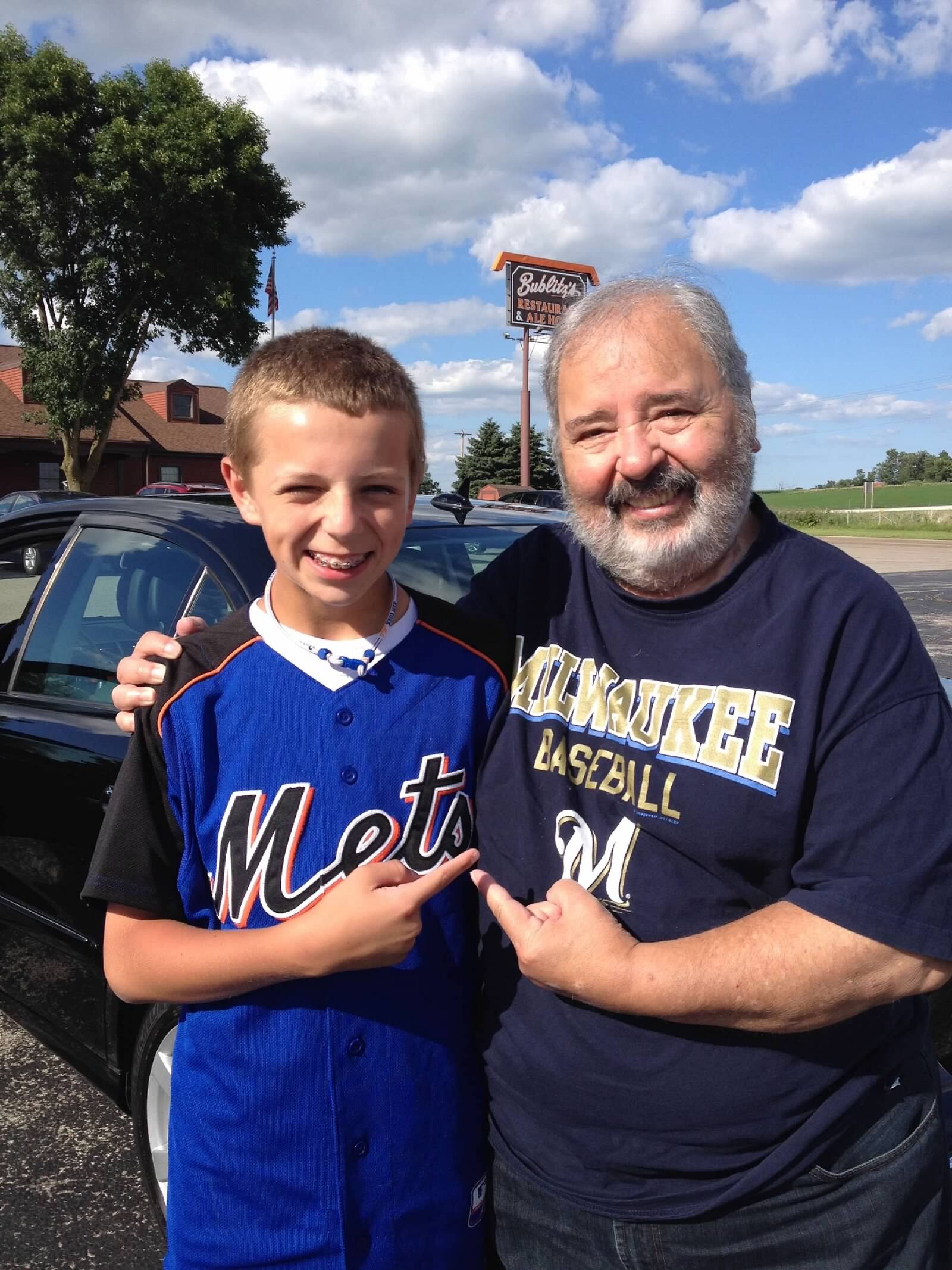 Two smiling males, a boy in a Mets jersey and an older man in a navy shirt, posing together outdoors in a parking lot.