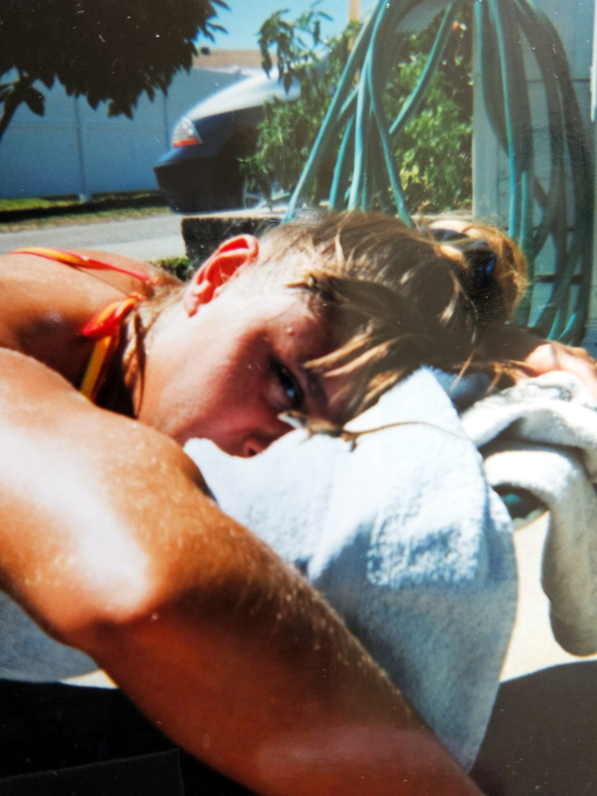 Person lying on a towel sunbathing outdoors, close-up of face and shoulder with a towel nearby; background shows a car and garden hose.