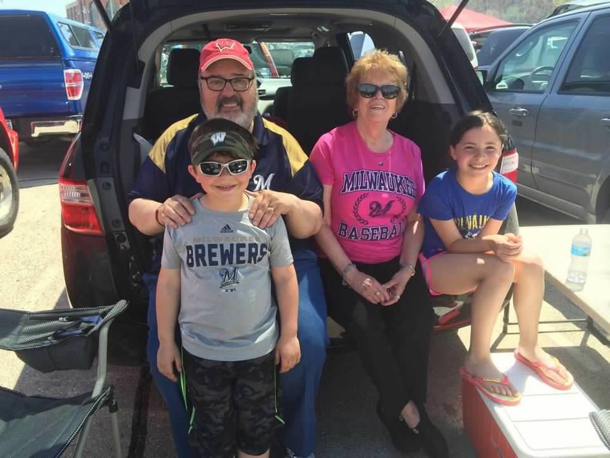 Family of four posing in the open SUV trunk at a parking lot (man in red cap, woman in pink shirt, two kids wearing team shirts).