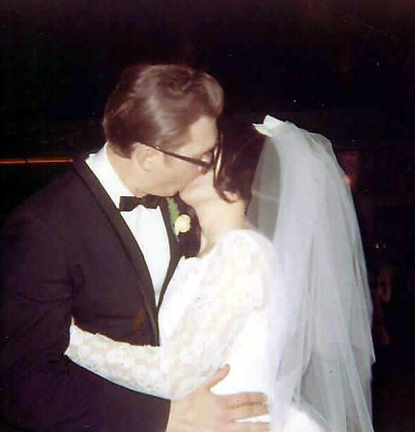 Bride and groom kiss at their wedding; the groom wears a black tuxedo and bow tie, the bride in a white lace gown with a veil.
