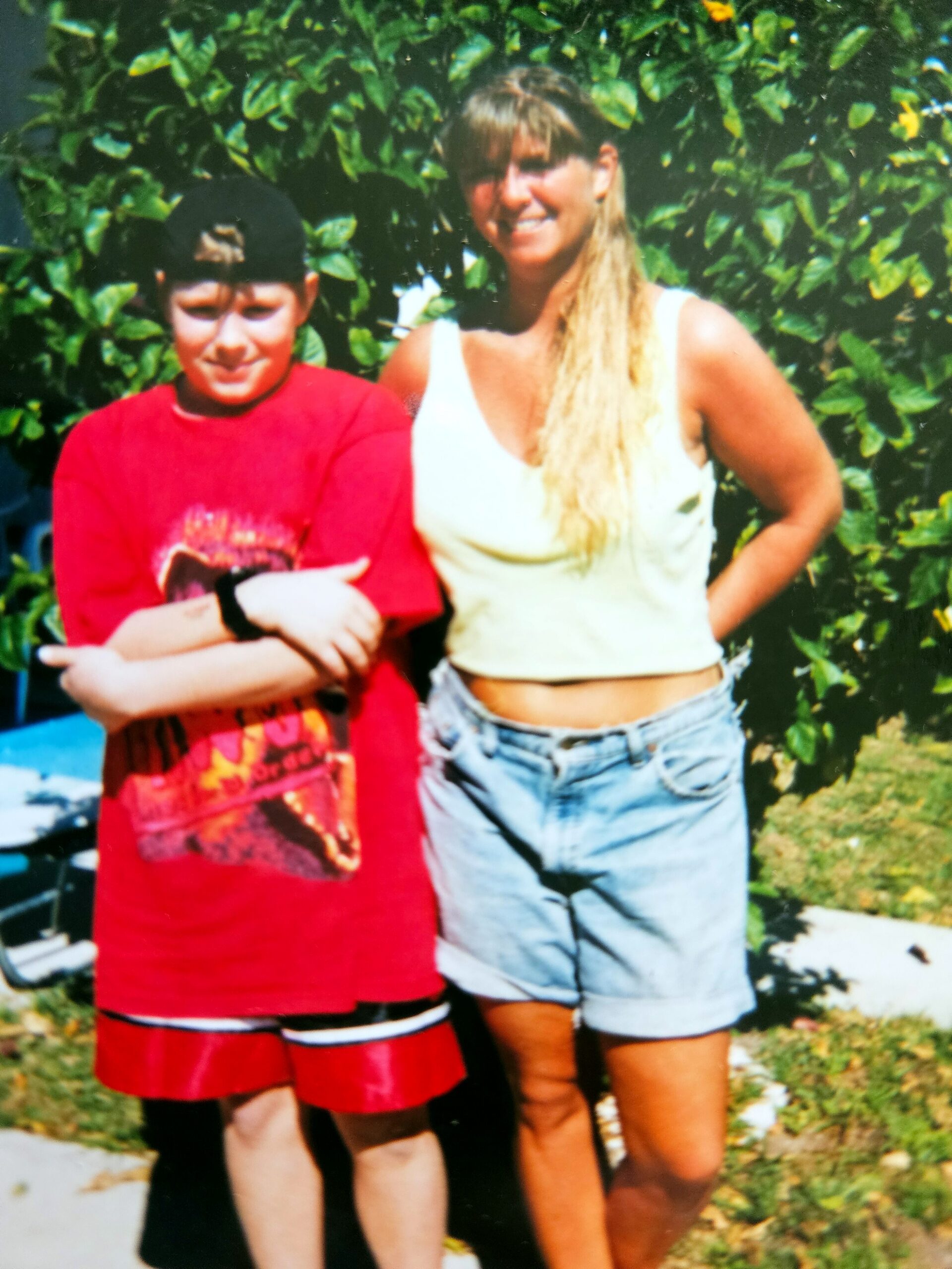 Two people posing outdoors in front of green bushes: a boy in a red shirt with folded arms and a woman in a white tank top smiling nearby.