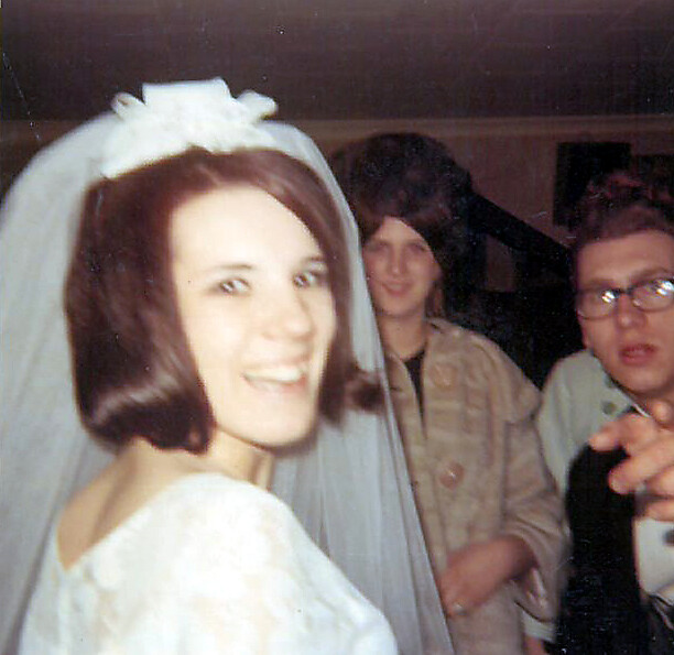 Bride in a white wedding dress and veil smiles at the camera, with two guests in the background.