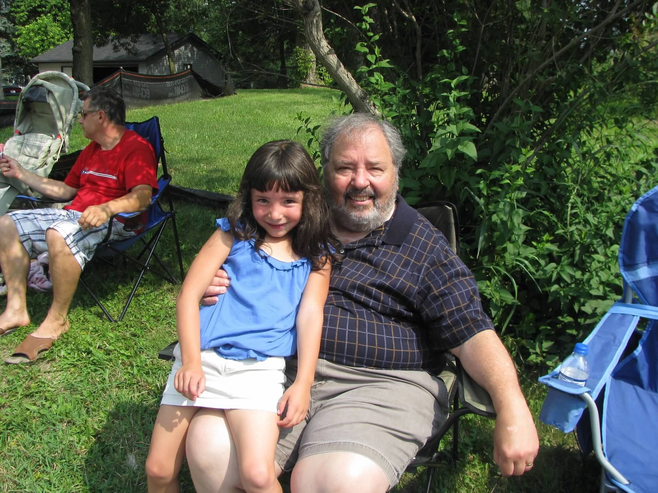 Grandfather with gray beard sits outdoors on a lawn, smiling beside young girl in blue top amid folding chairs and greenery.