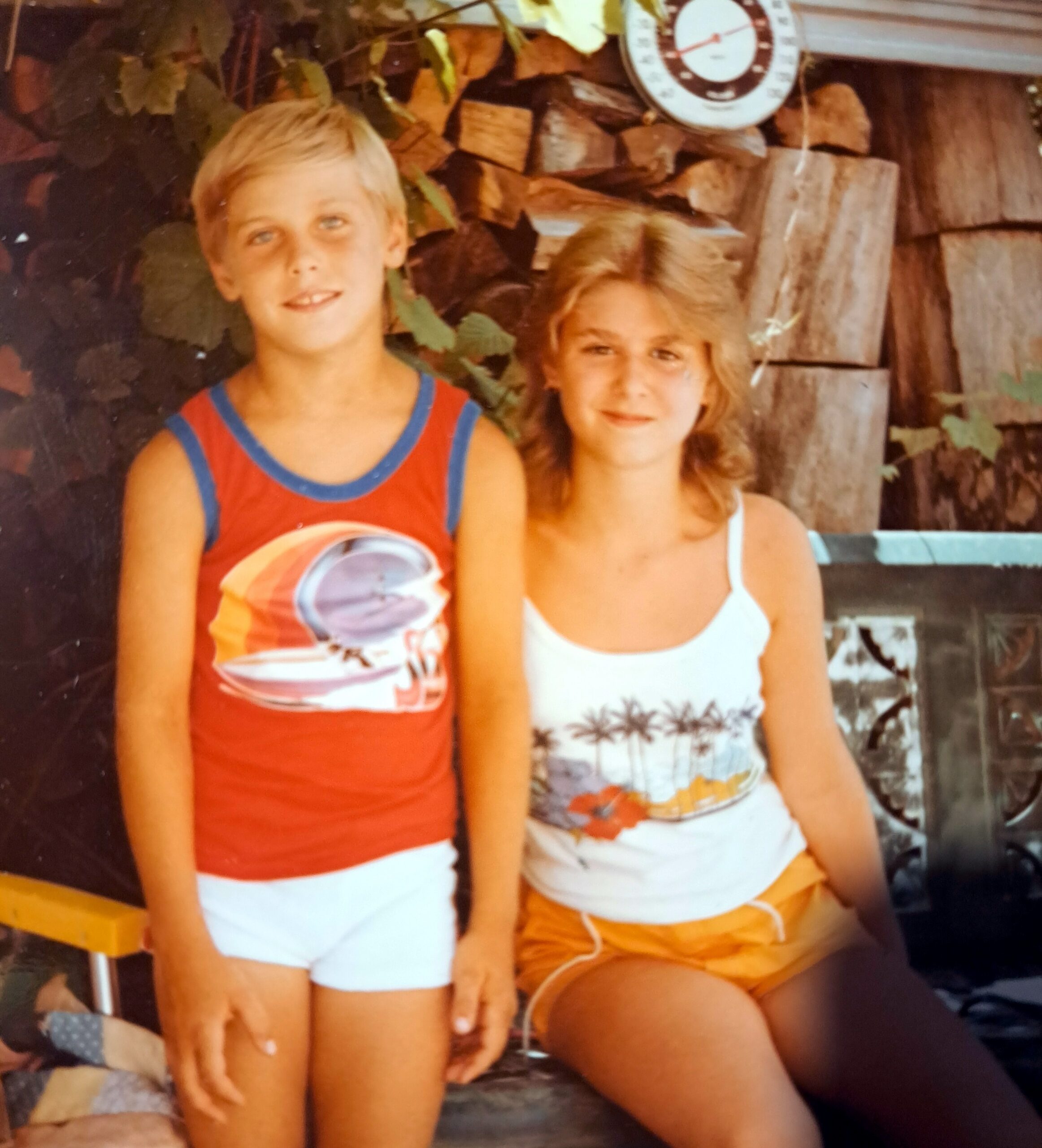 Two kids posing together in summer outfits: a blond boy in a red tank top and white shorts and a girl in a white top with palm-printed shorts, standing in front of stacked logs.