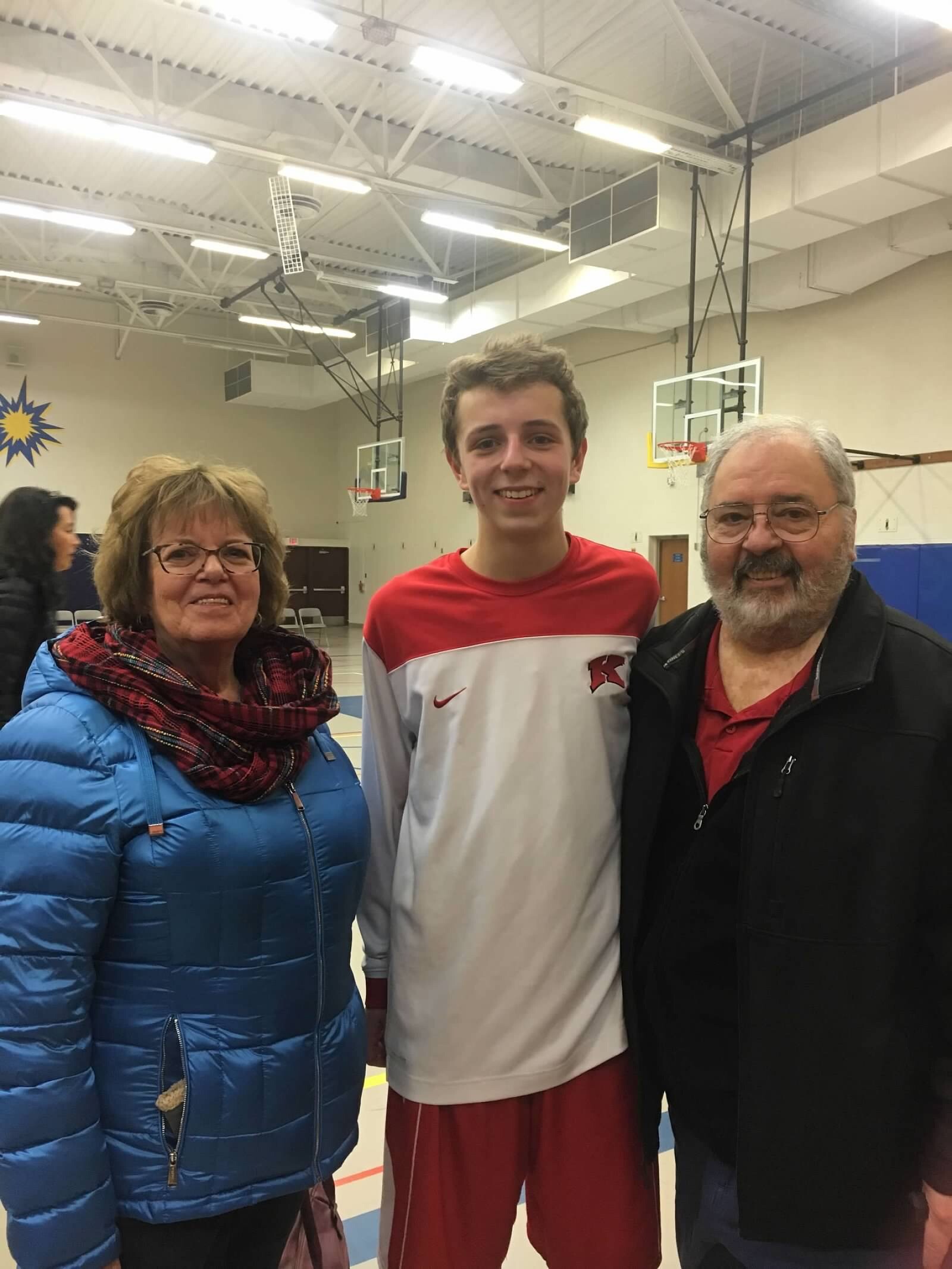 Three people posing for a photo in a school gym: a woman in a blue puffer jacket, a teenage boy in red and white athletic wear, and an older man in a black jacket nearby.
