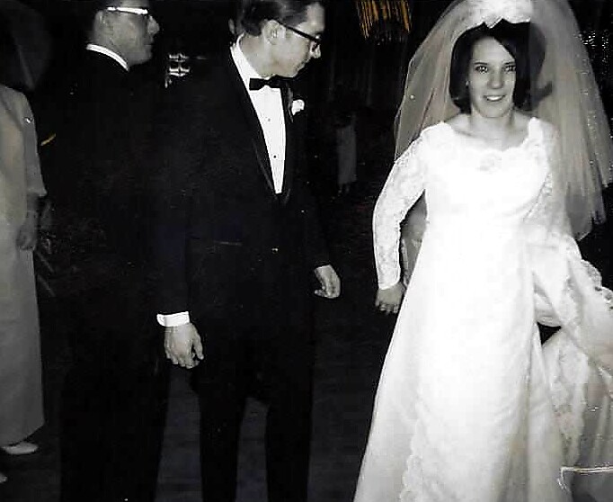 Bride in a white wedding gown and veil holds hands with a man in a black tuxedo at their wedding.
