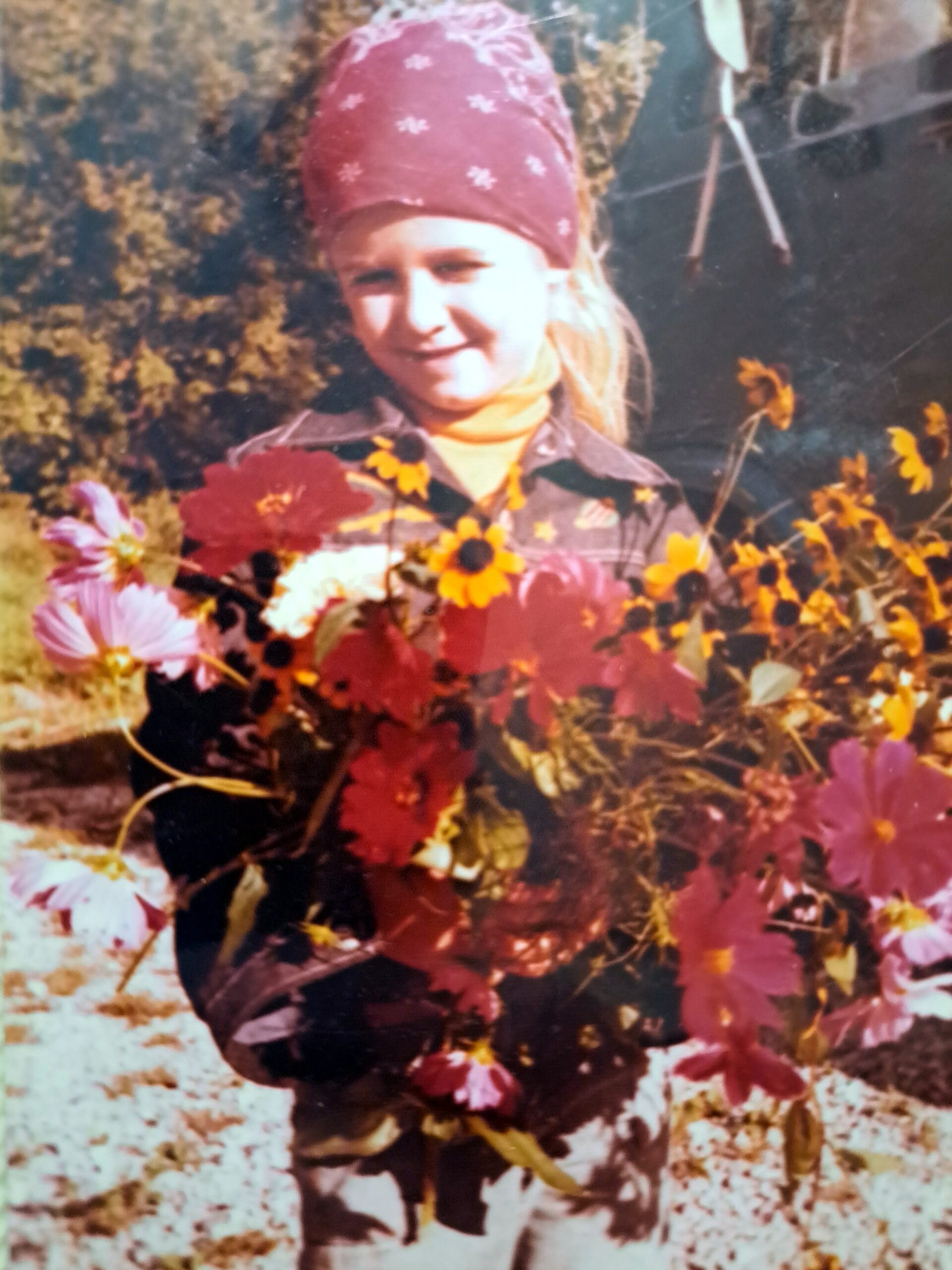 Person wearing a red bandana and brown jacket, holding a bright bouquet of autumn flowers outdoors in warm light.