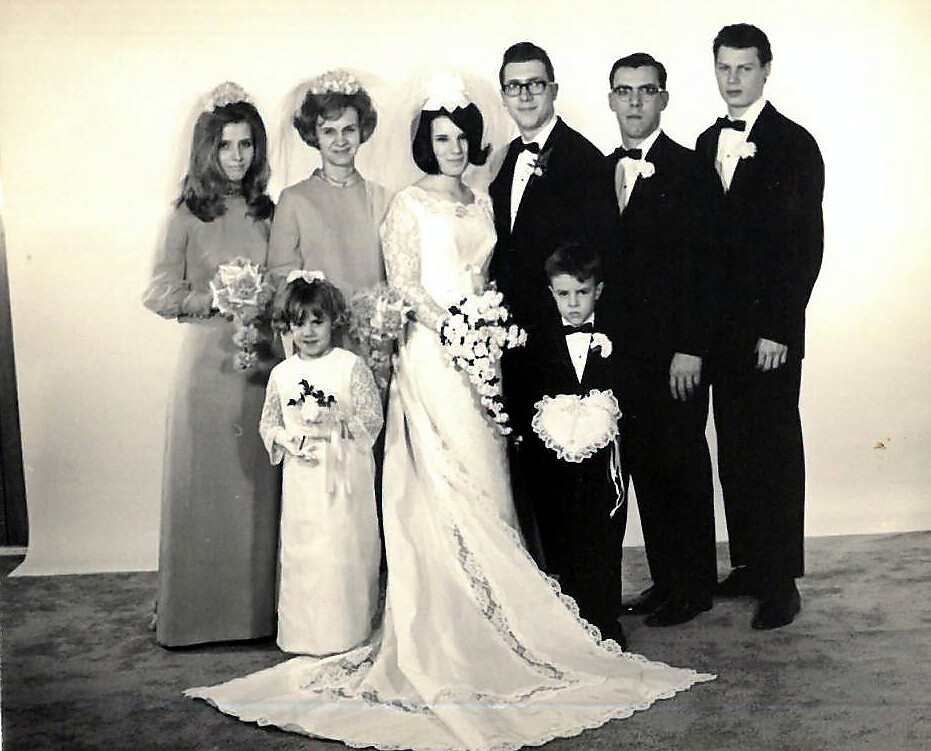 Bride in a white gown with veil holding a bouquet, flanked by the wedding party in formal attire including men in tuxedos and two girls in white dresses with bouquets.