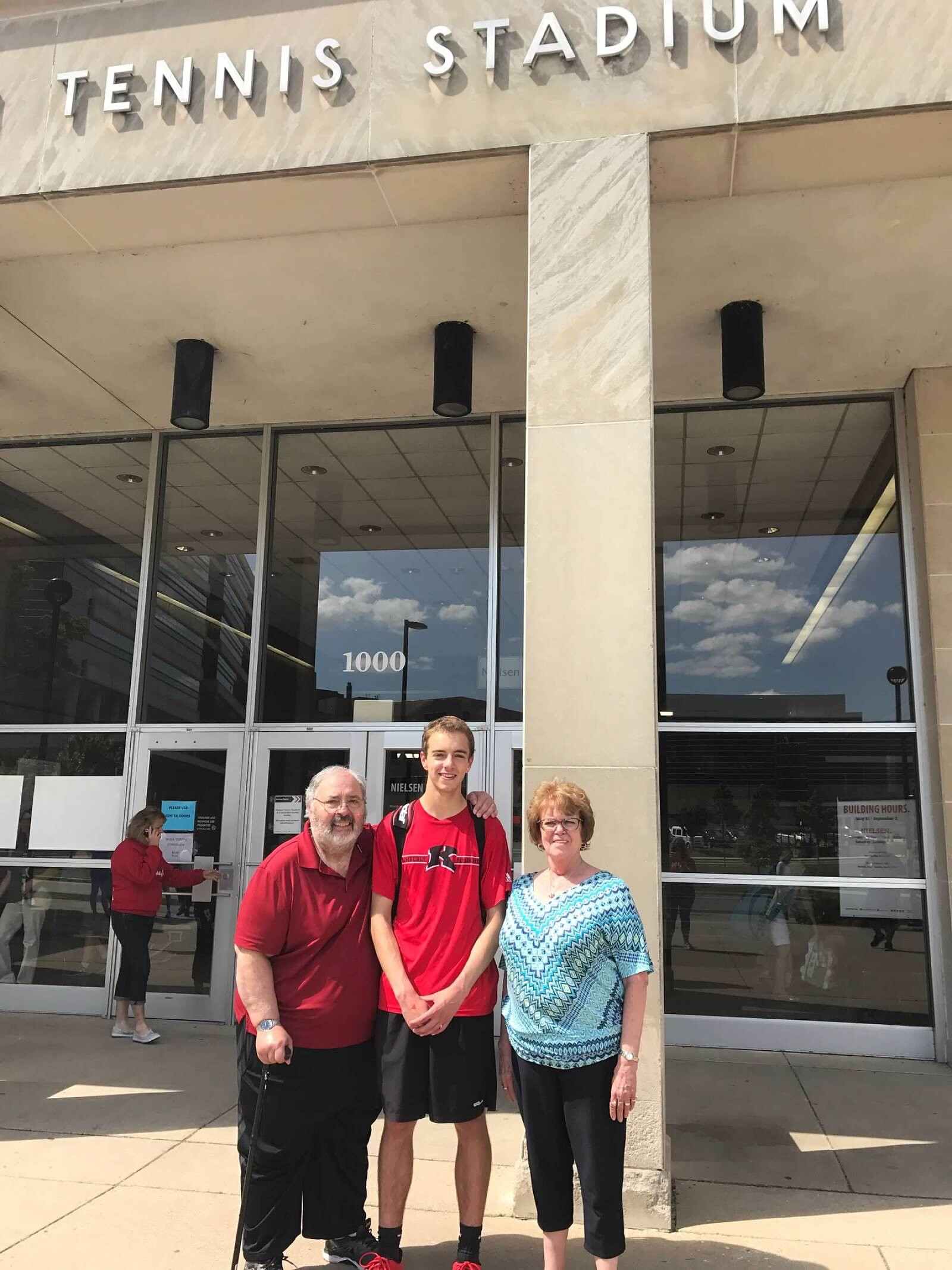 Three people stand together outside a tennis stadium entrance, smiling, with 'TENNIS STADIUM' visible above.