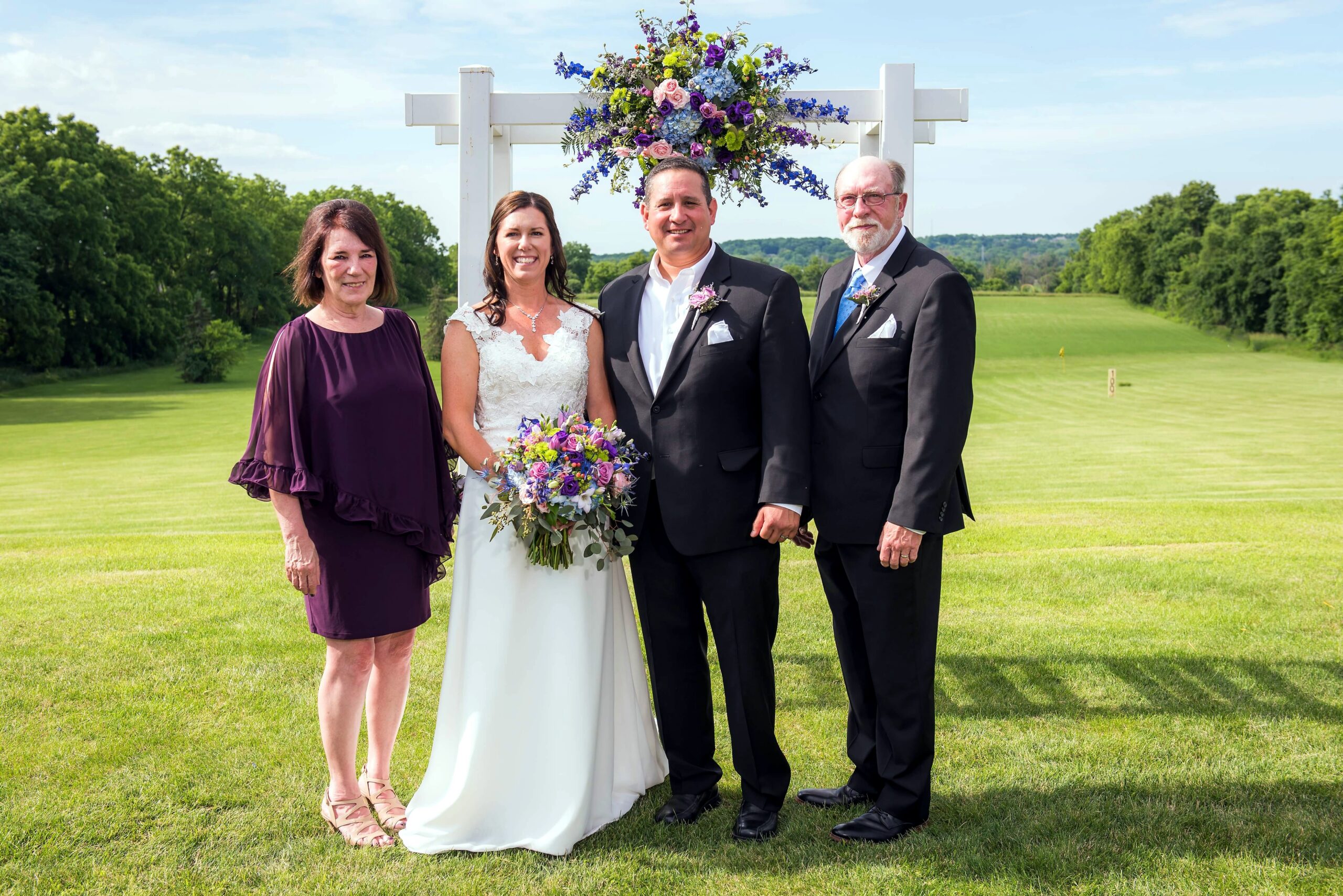 Bride in a white wedding gown holding a colorful bouquet, flanked by two men in black suits and a woman in a purple dress, outdoors under a floral arch on a sunny day.