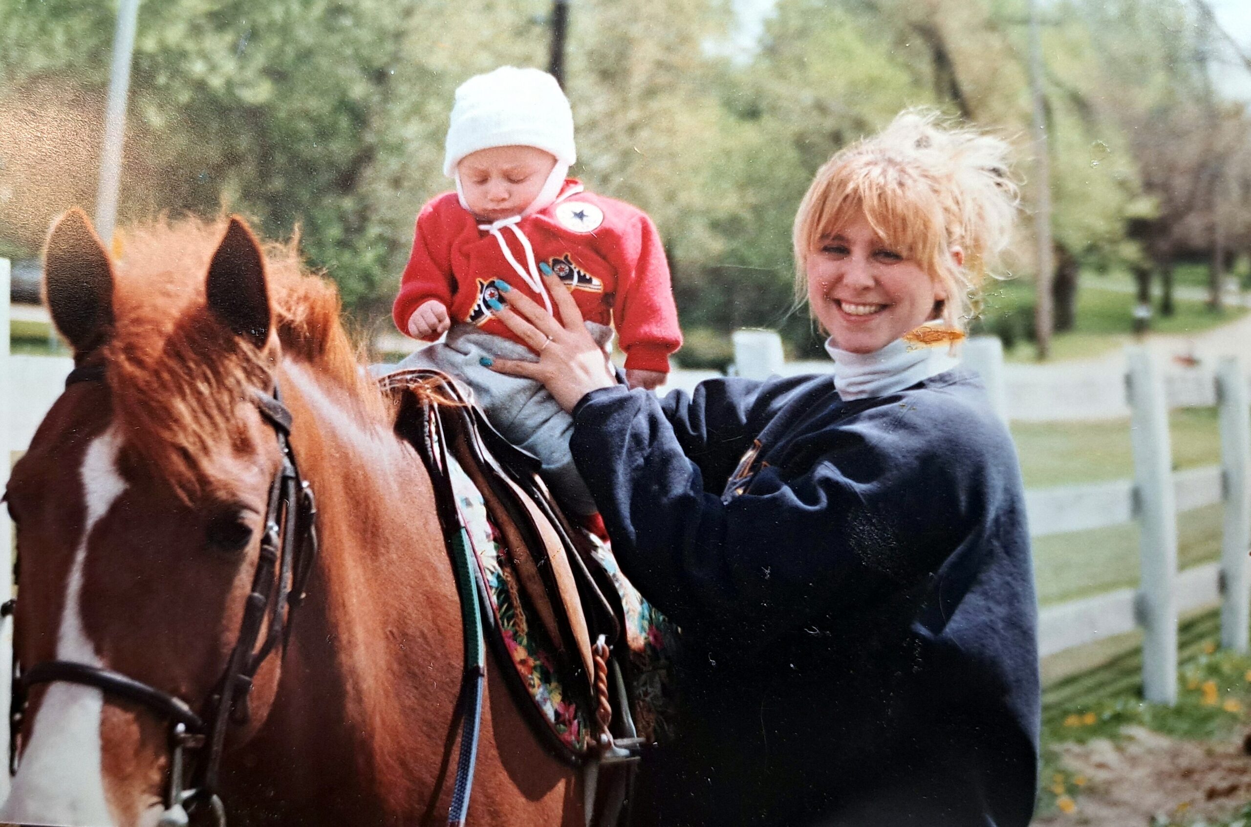Smiling woman on a brown horse lifts a baby in a white hat and red jacket outdoors by a fence and trees.