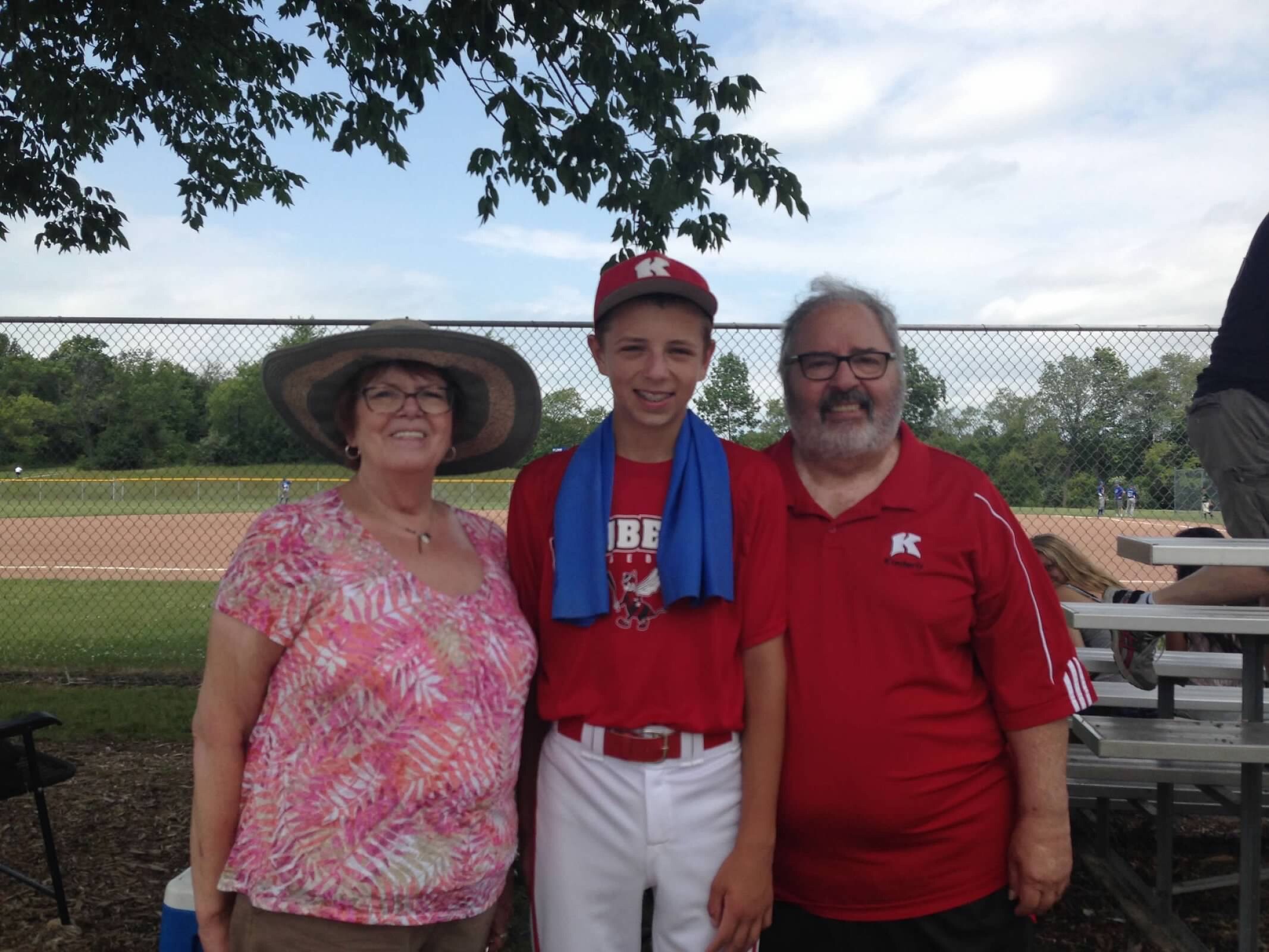 Three people posing together at a baseball field: an older woman in a wide hat, a young man in a red cap and shirt with a blue towel, and an older man in a red polo.