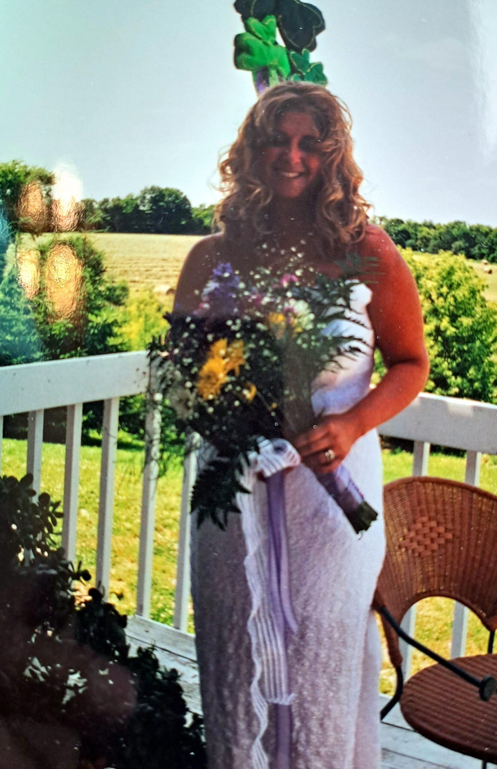Smiling woman in a white dress holds a colorful bouquet outdoors on a wooden deck railing.