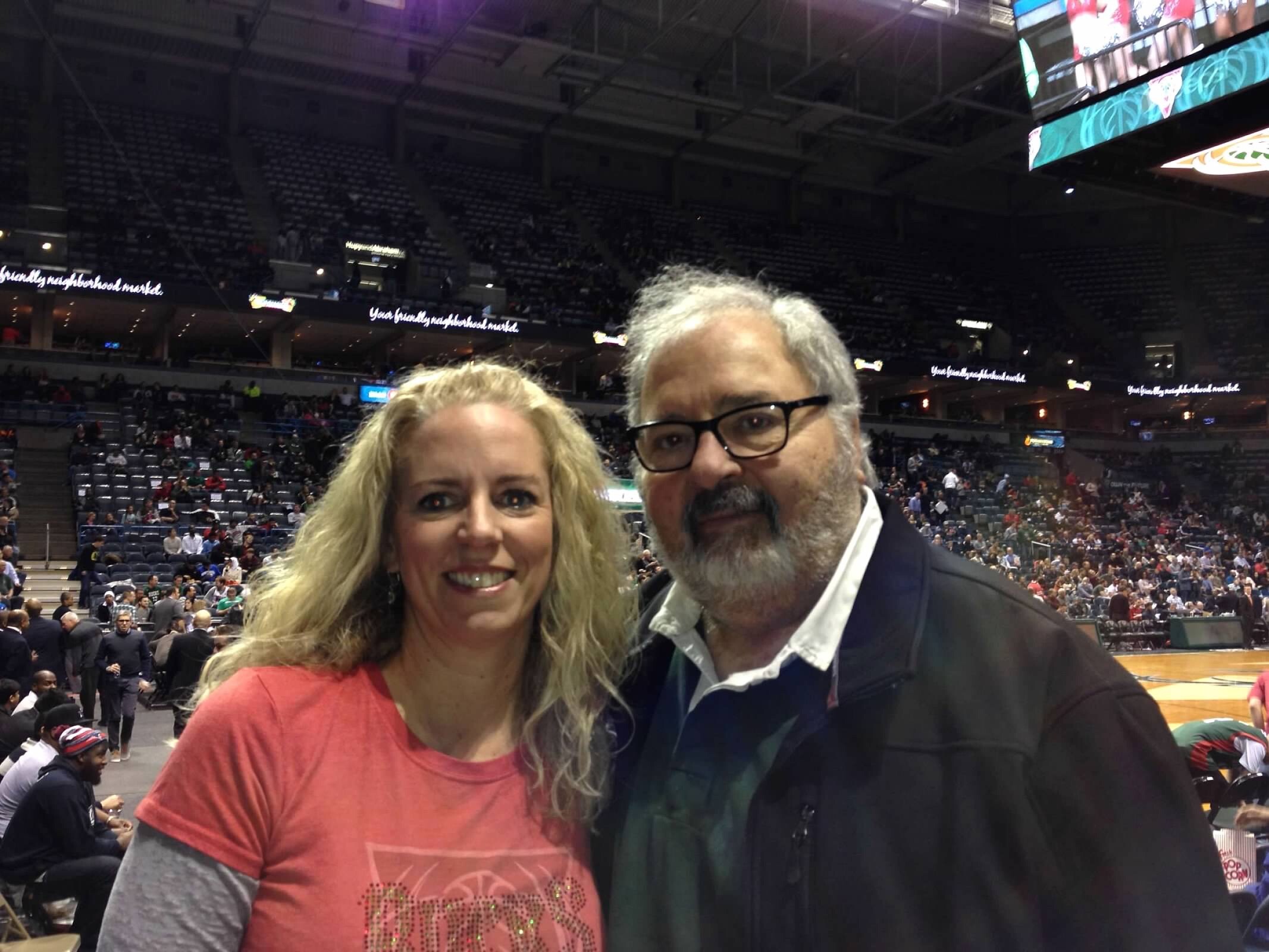 Two adults taking a selfie in a large indoor sports arena with bleachers and a crowd in the background.