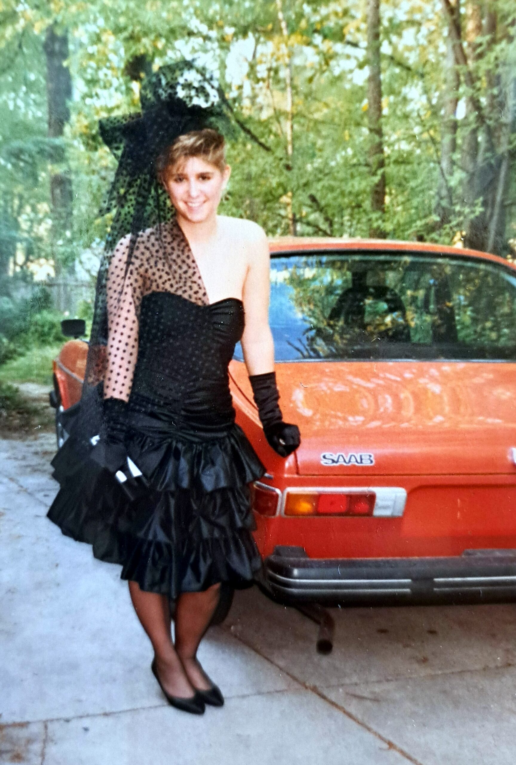 Woman in a black, strapless evening gown with a dotted sheer overlay stands beside an orange Saab car outdoors amidst trees.