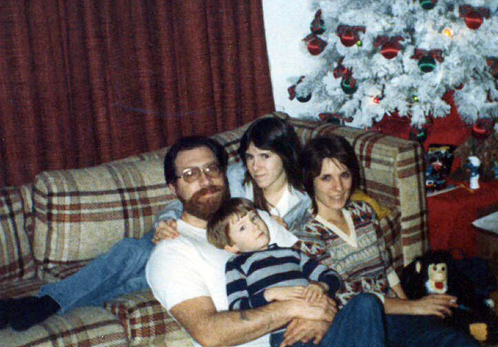 Family of four sits on a plaid couch in a living room, with a white Christmas tree in the background.