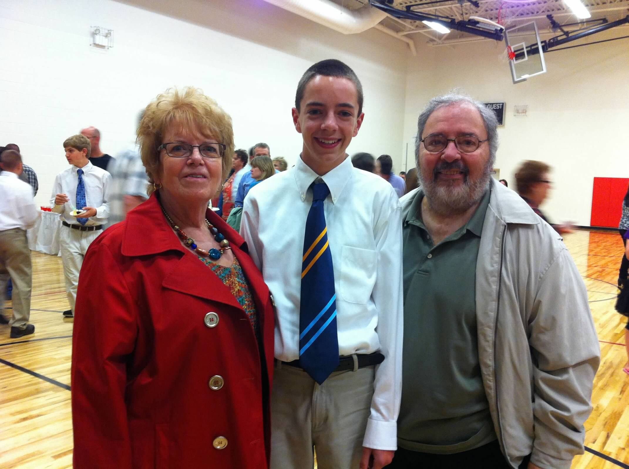 Three people pose for a photo in a gym: woman in a red coat, a young man in a white shirt and blue striped tie, and an older man with a gray beard smiling at the camera.