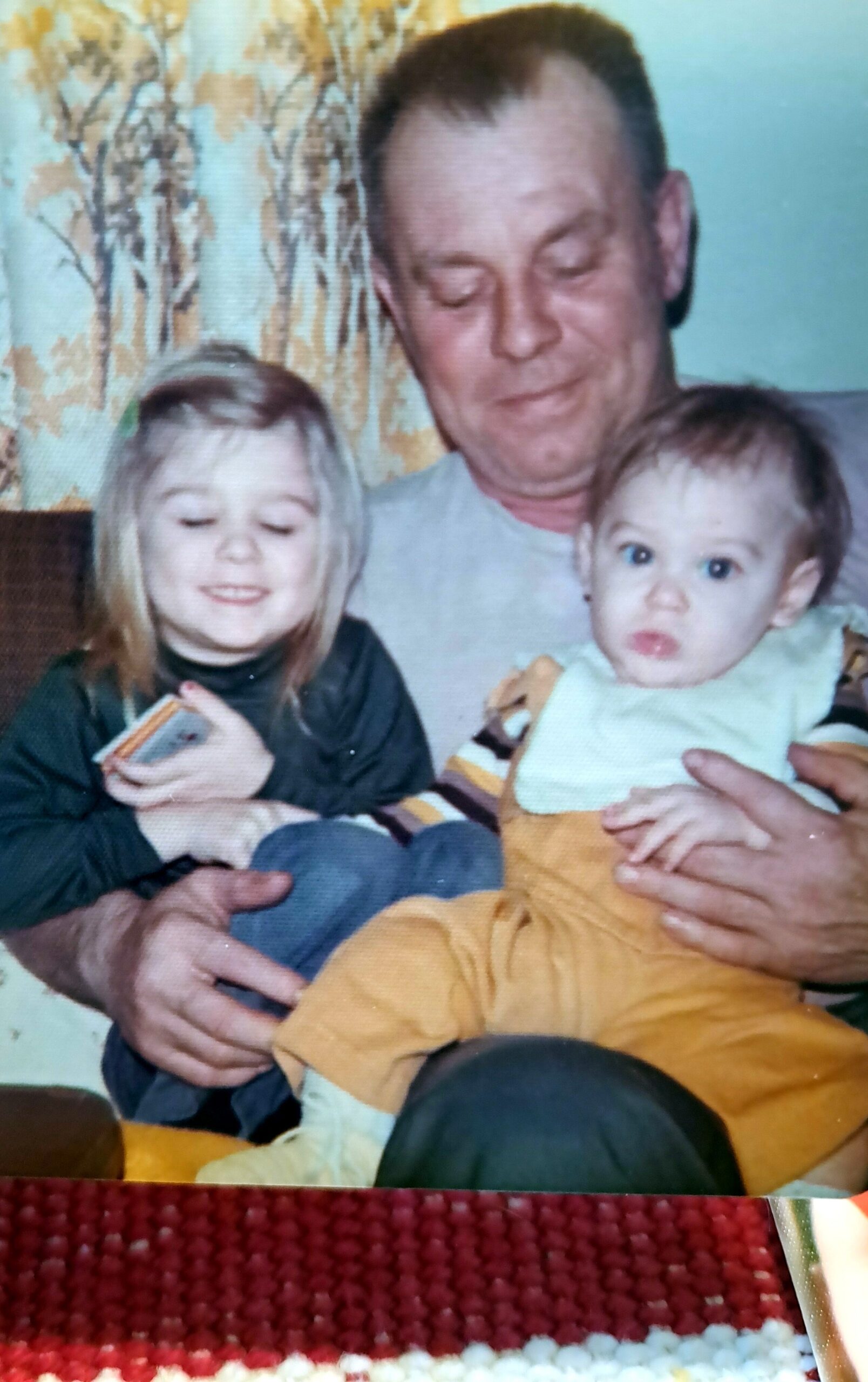 Man sitting with two small children: a smiling blonde girl on his left and a baby with blue eyes on his right in his arms.