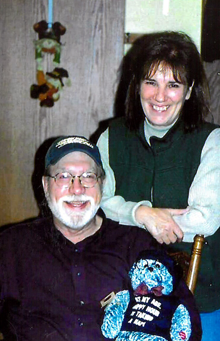 Smiling older man with a white beard and glasses sits in a dark shirt and cap; a smiling woman stands behind him with her arm on the chair, and a blue teddy bear rests in front with a printed shirt.