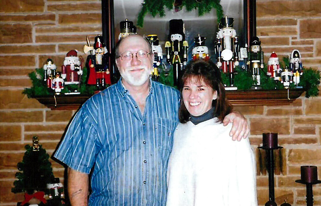 Smiling couple posing arm-in-arm in front of a brick fireplace decorated with nutcracker figurines on the mantle.