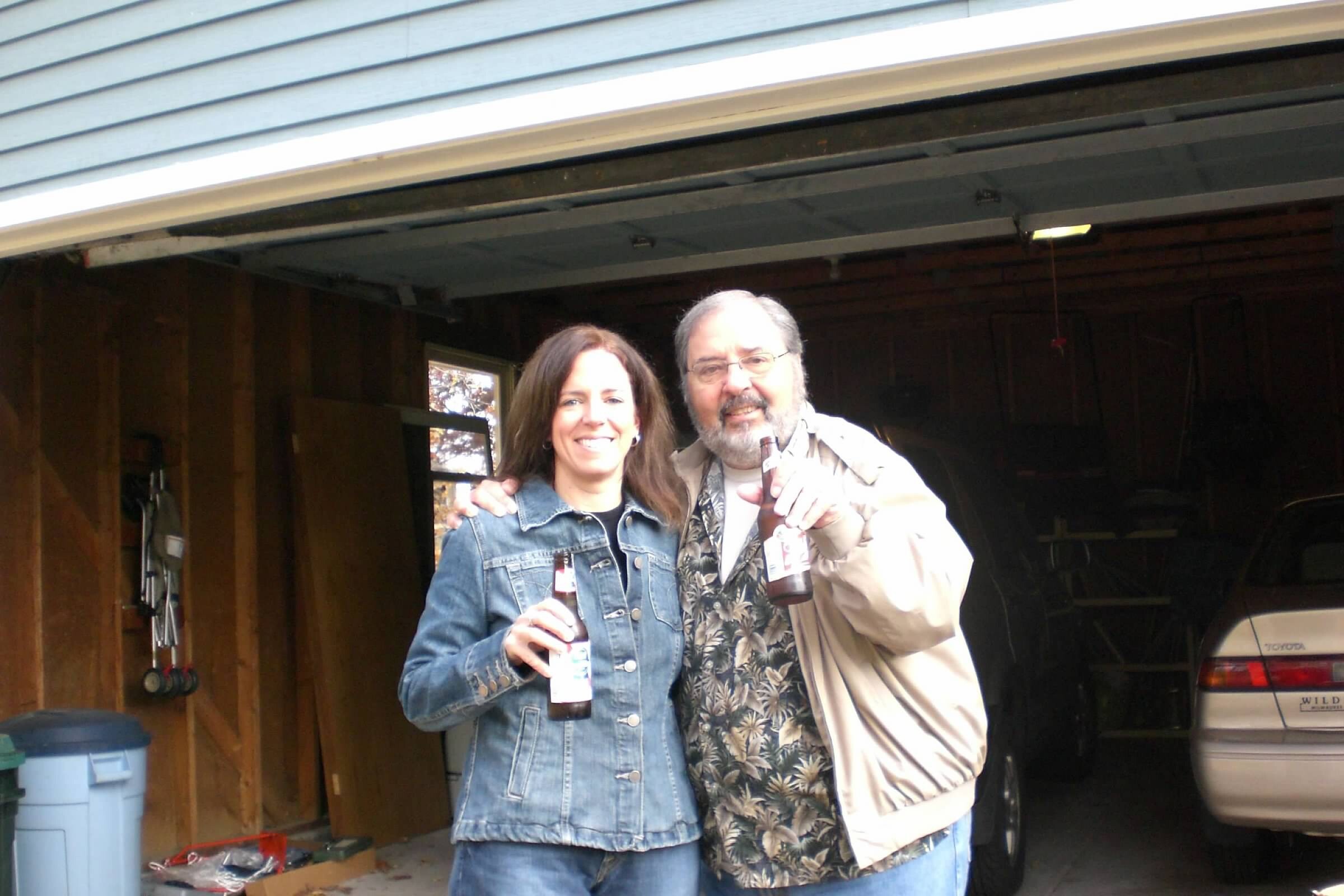 Two adults posing in a garage, each holding a beer and smiling at the camera.