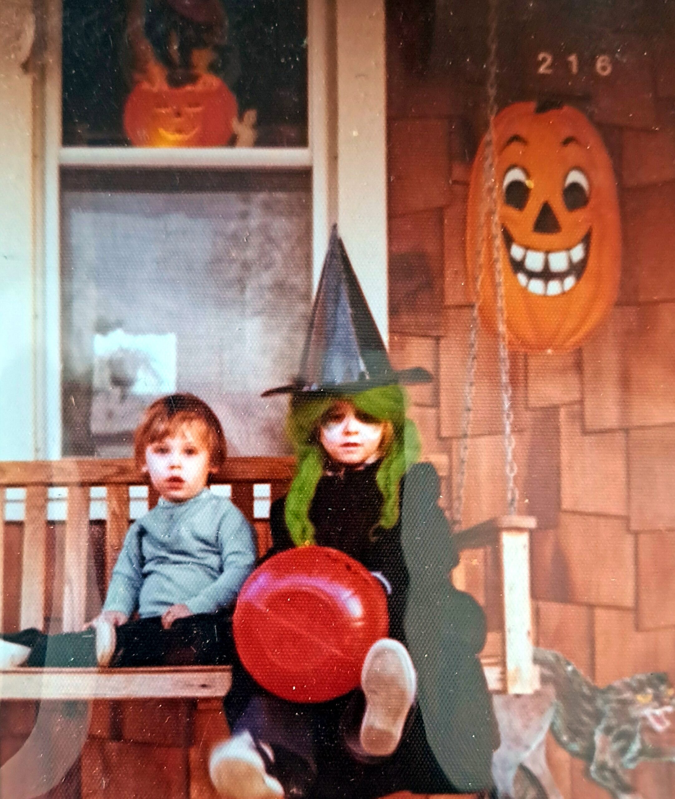 Two children sit on a porch with Halloween decorations, including a large jack-o'-lantern prop on the right.