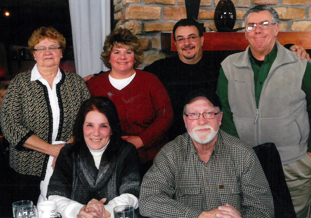 Group of six adults smiling for a photo in a living room with a stone fireplace in the background; three seated at a table and three standing behind them.