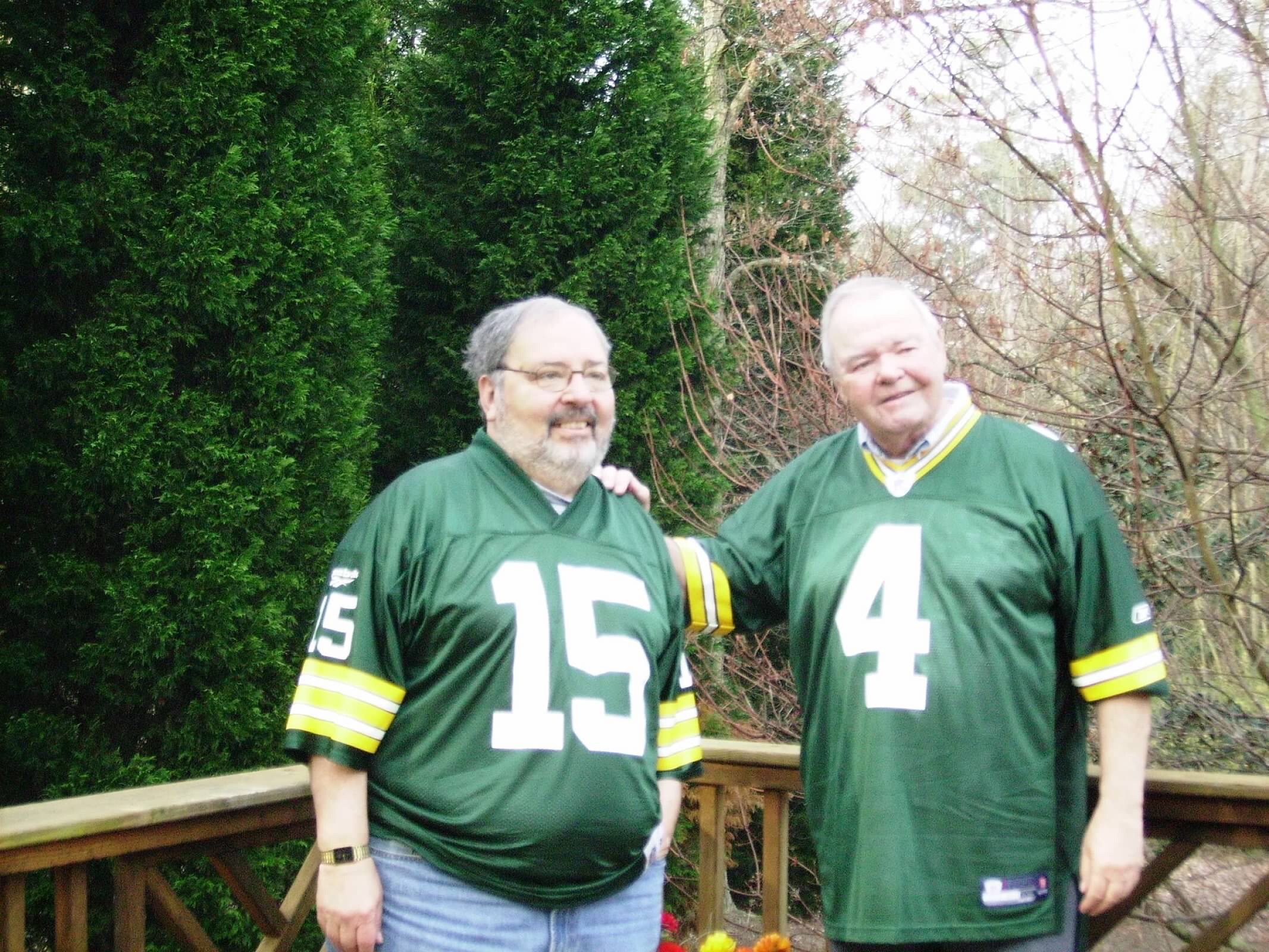 Two men in green jerseys with white numbers 15 and 14 pose outdoors, smiling with a wooden railing behind them and evergreen trees nearby.