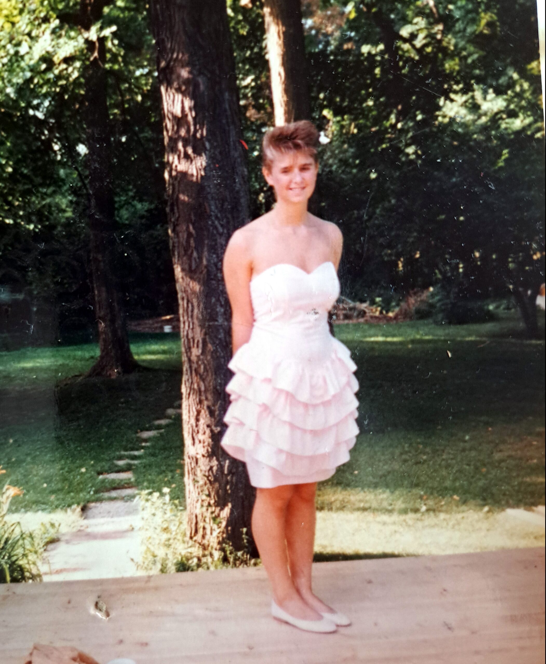 Young woman in a light pink strapless dress with layered ruffles, standing next to a tree in a park on a sunny day.