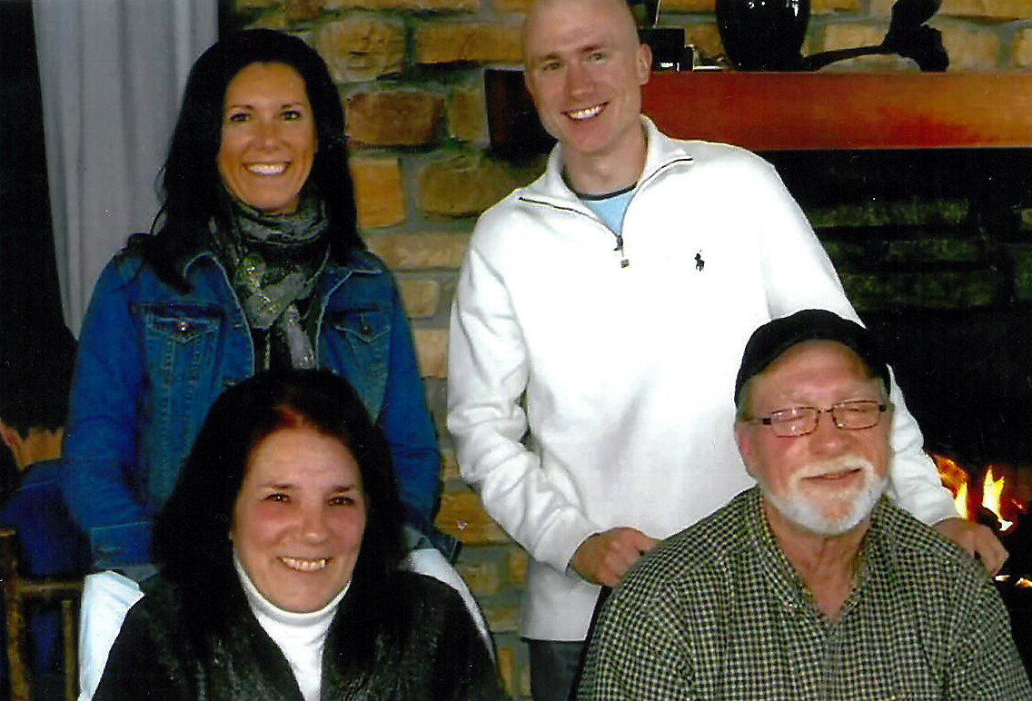 Four adults posing indoors by a stone fireplace, smiling at the camera.