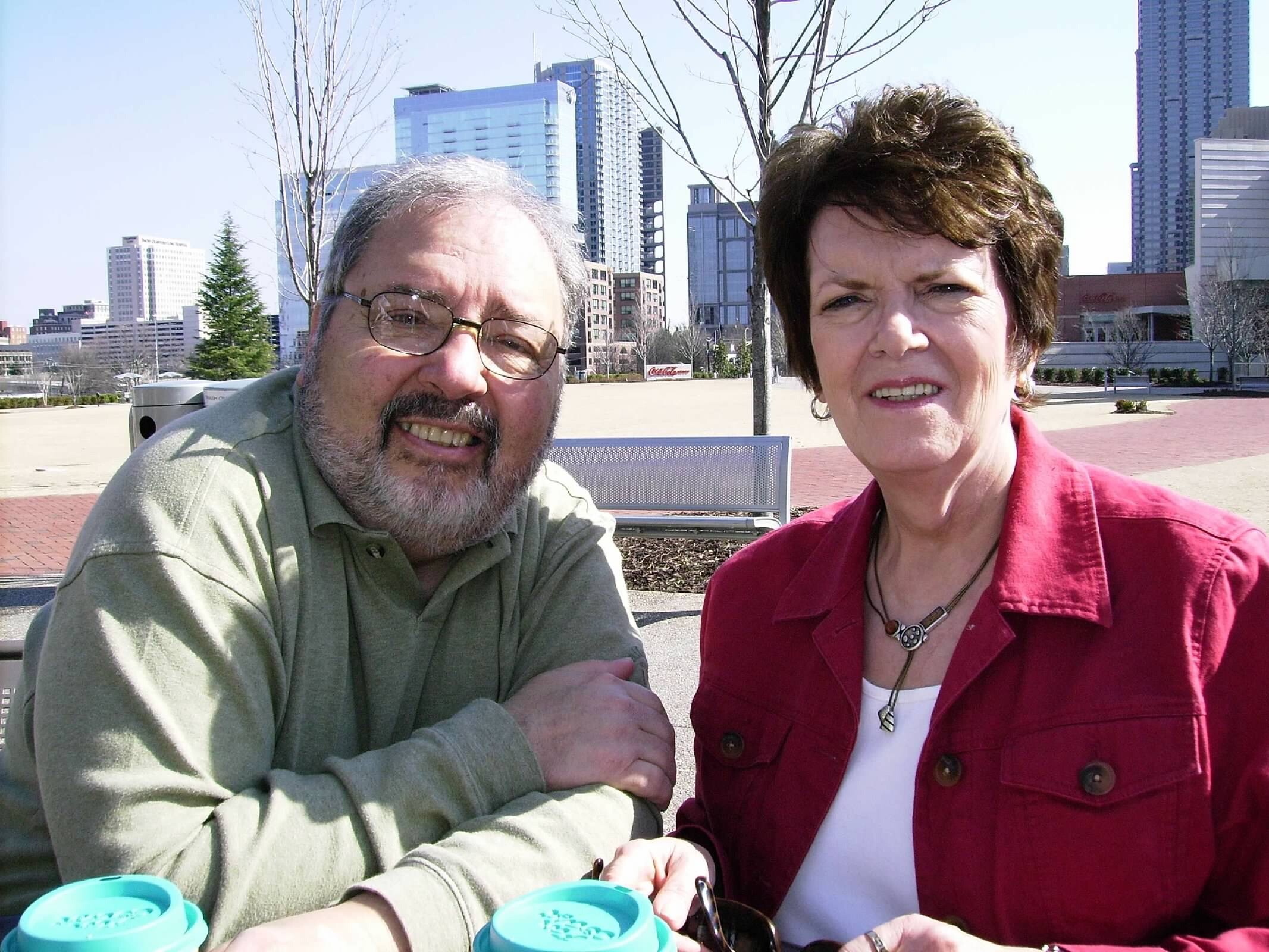 Smiling man with glasses sits next to a woman in a red jacket, outdoor plaza with skyscrapers in the background.