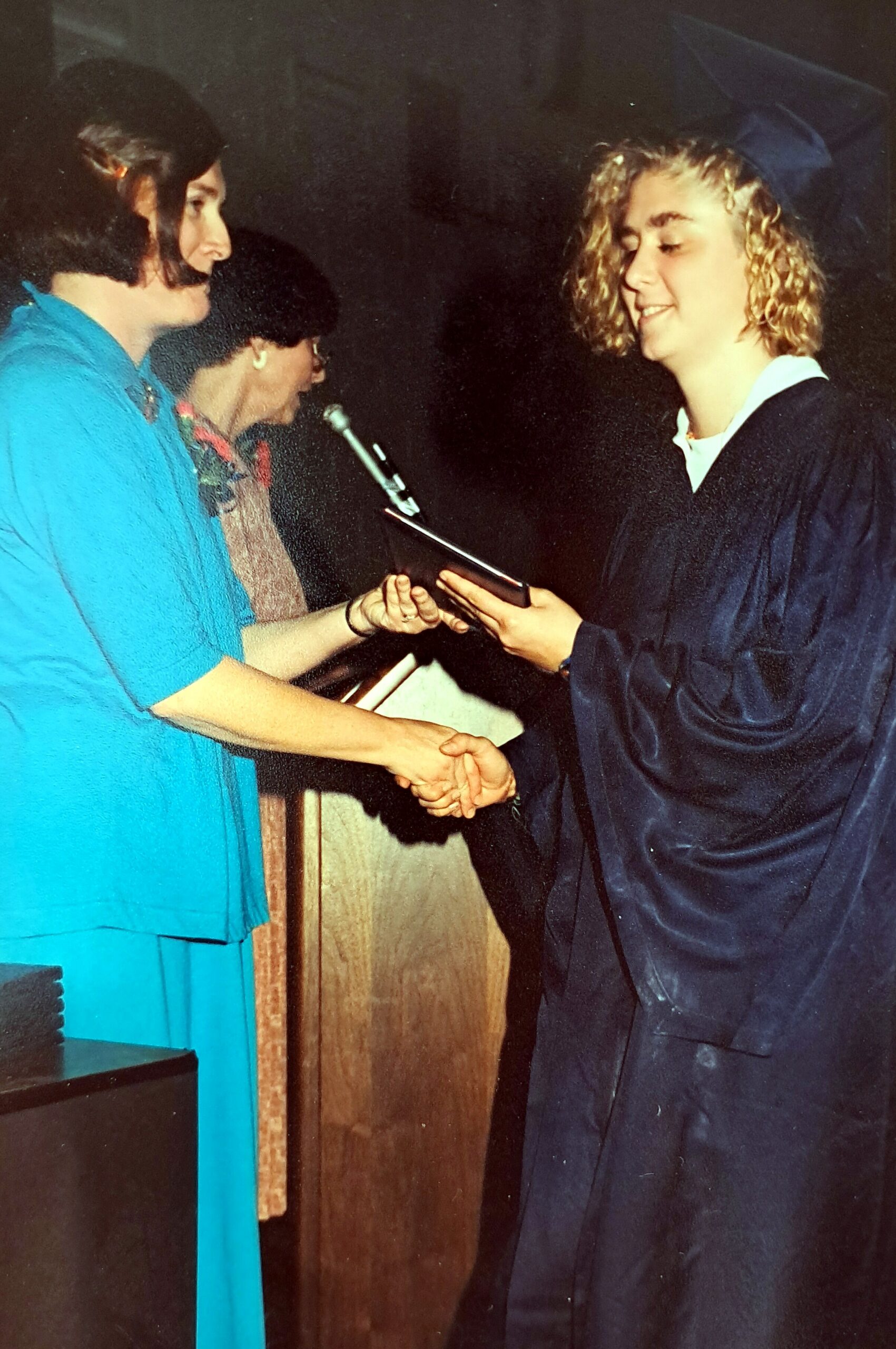 Graduate in cap and gown shakes hands with a woman in a teal outfit during a ceremony, holding a folder/diploma slot
