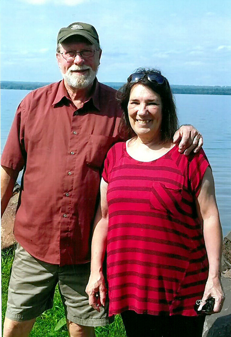 Older couple standing together by a lake, the man with a cap and beard has his arm around the woman, smiling at the camera.