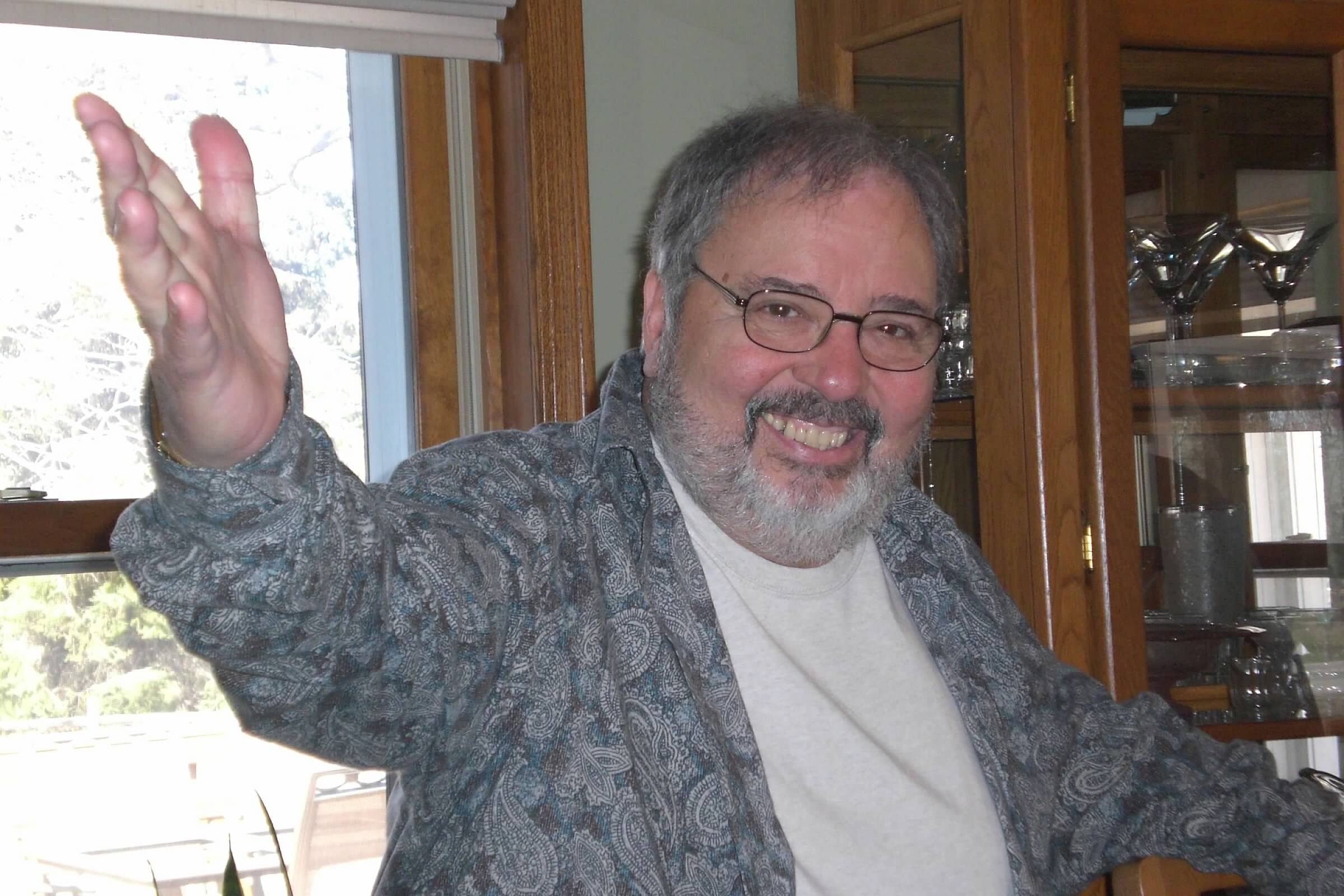 Smiling man with gray hair and glasses waving hello indoors.