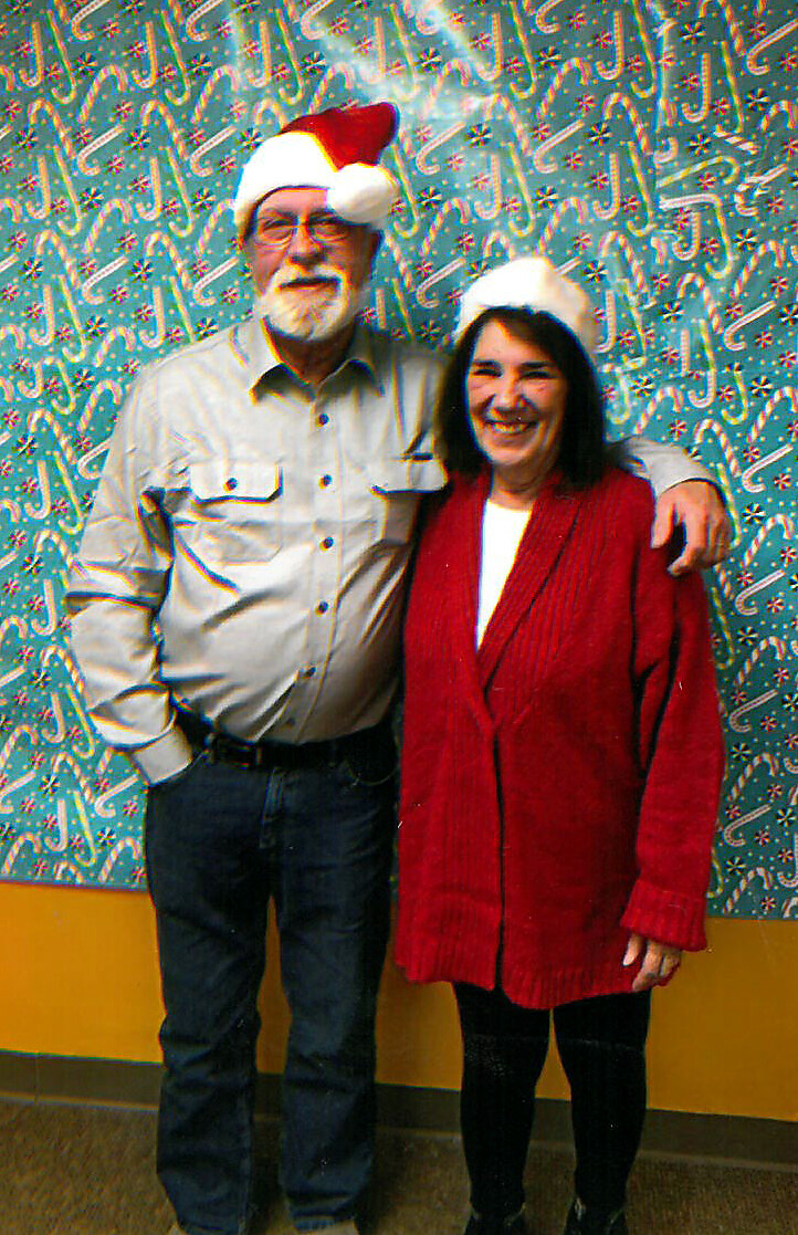 Older couple wearing Santa hats pose together and smile in front of festive patterned wallpaper.