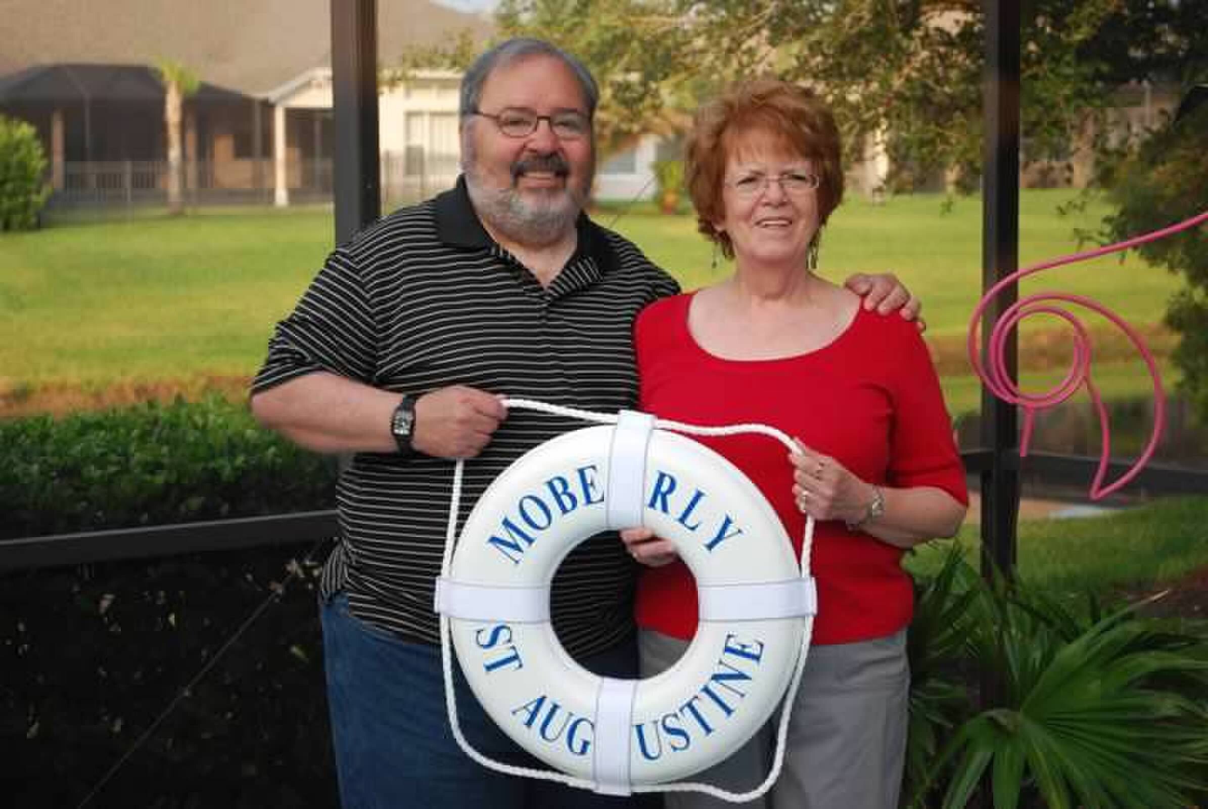 Smiling couple outdoors holding a white lifebuoy with blue lettering.