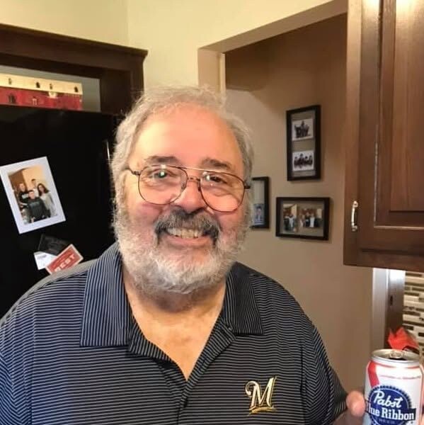 Smiling older man with gray hair and beard, wearing glasses and a striped polo with an M logo, standing in a kitchen.