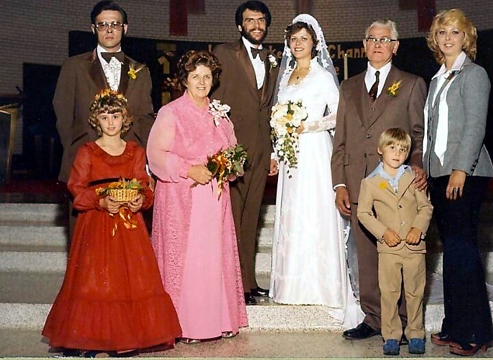Wedding party on church steps: bride in white gown with bouquet, groom in brown suit, flanked by relatives in formal attire, plus a young girl in red dress with a basket and a boy in a tan suit.
