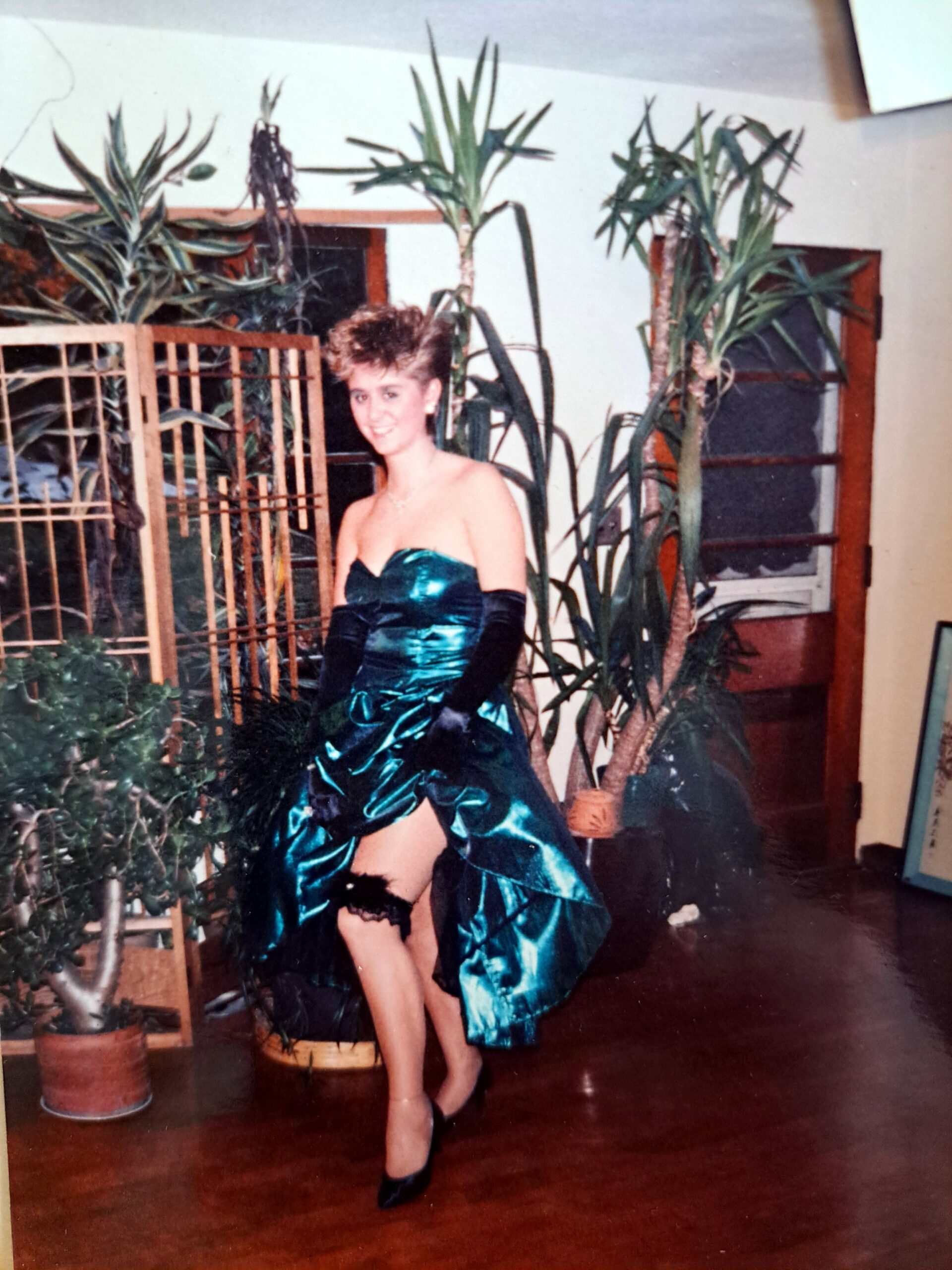 Smiling woman in a teal satin strapless gown with long black gloves posing among tall indoor plants and a wooden room divider.