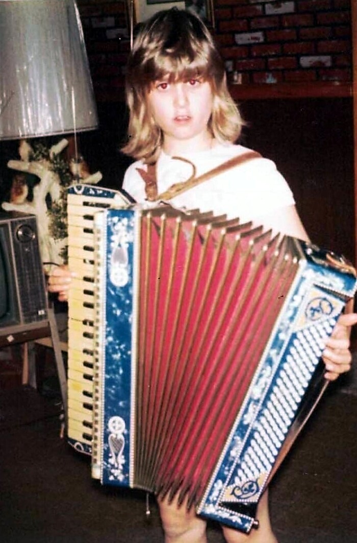 Child with shoulder-length hair playing a blue accordion indoors.