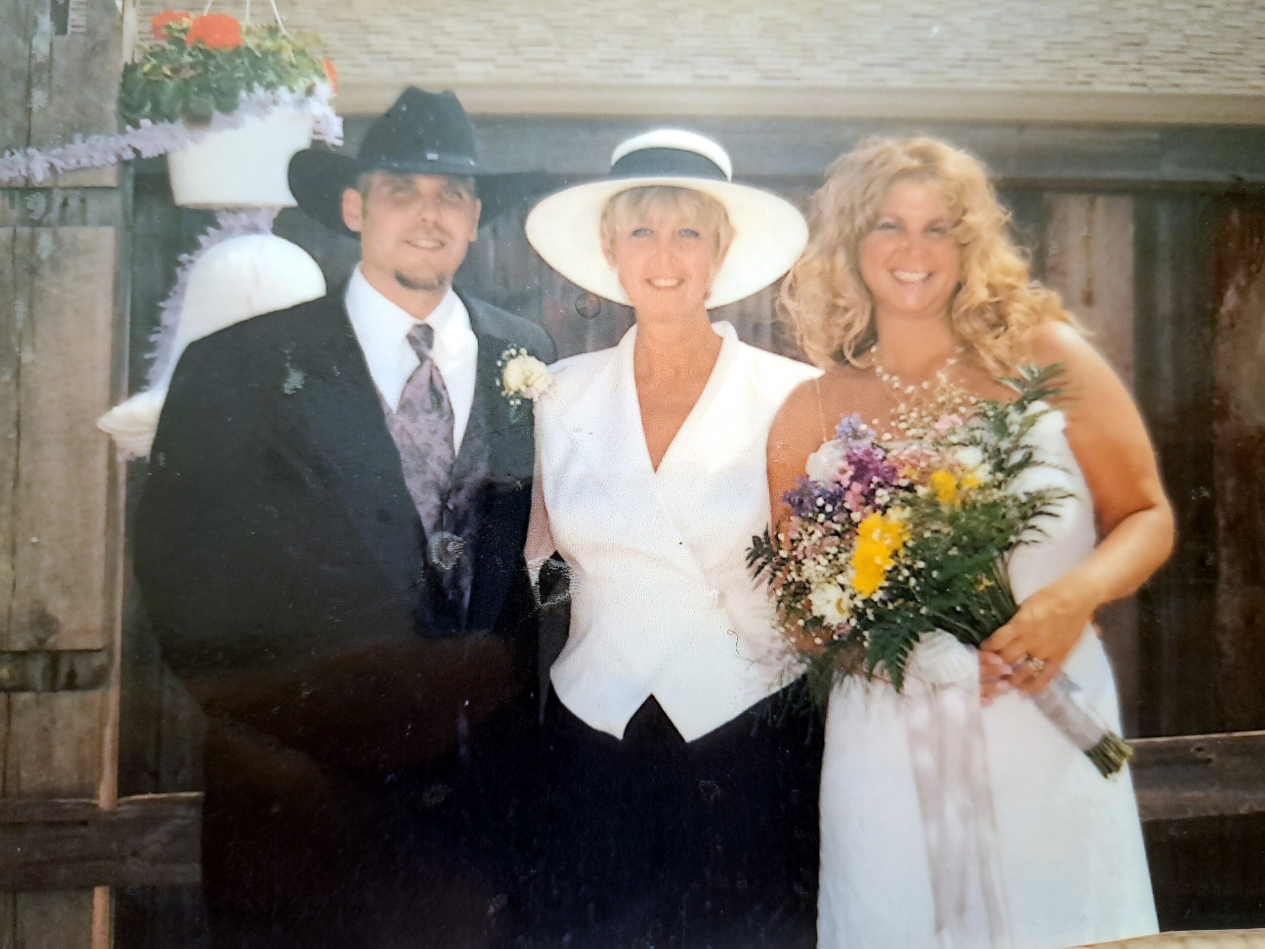 Three people at a wedding: a groom in a dark suit beside a bride in a white dress with a wide-brim hat, and a bridesmaid with a colorful bouquet.