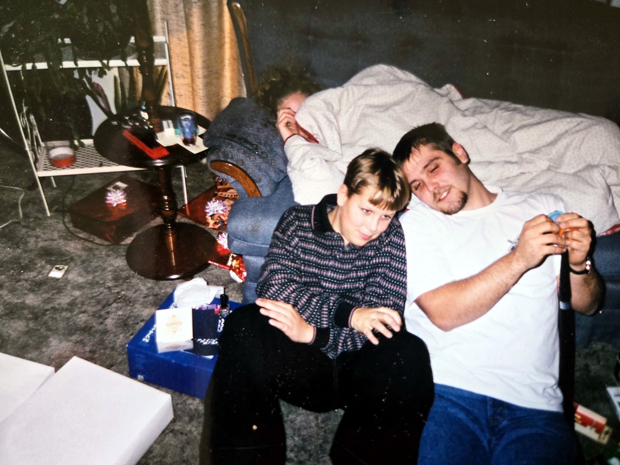 Two people lounging on the floor of a living room: a man in a white shirt and blanket lying down, a boy in a plaid shirt sits nearby amid clutter and presents.