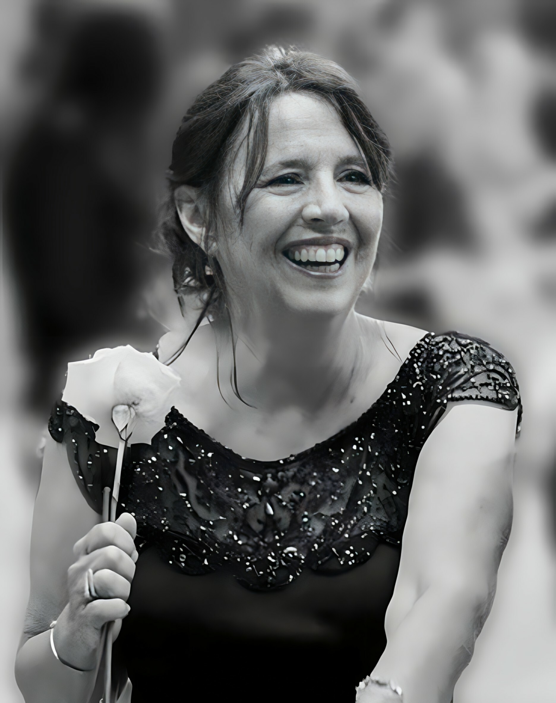 Woman smiling and laughing while holding a microphone stand, wearing a black sequined dress at a celebration; blurred crowd in background