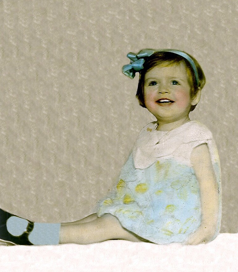 Smiling toddler girl in a pastel blue and white dress with a blue headband, sitting against a light backdrop.