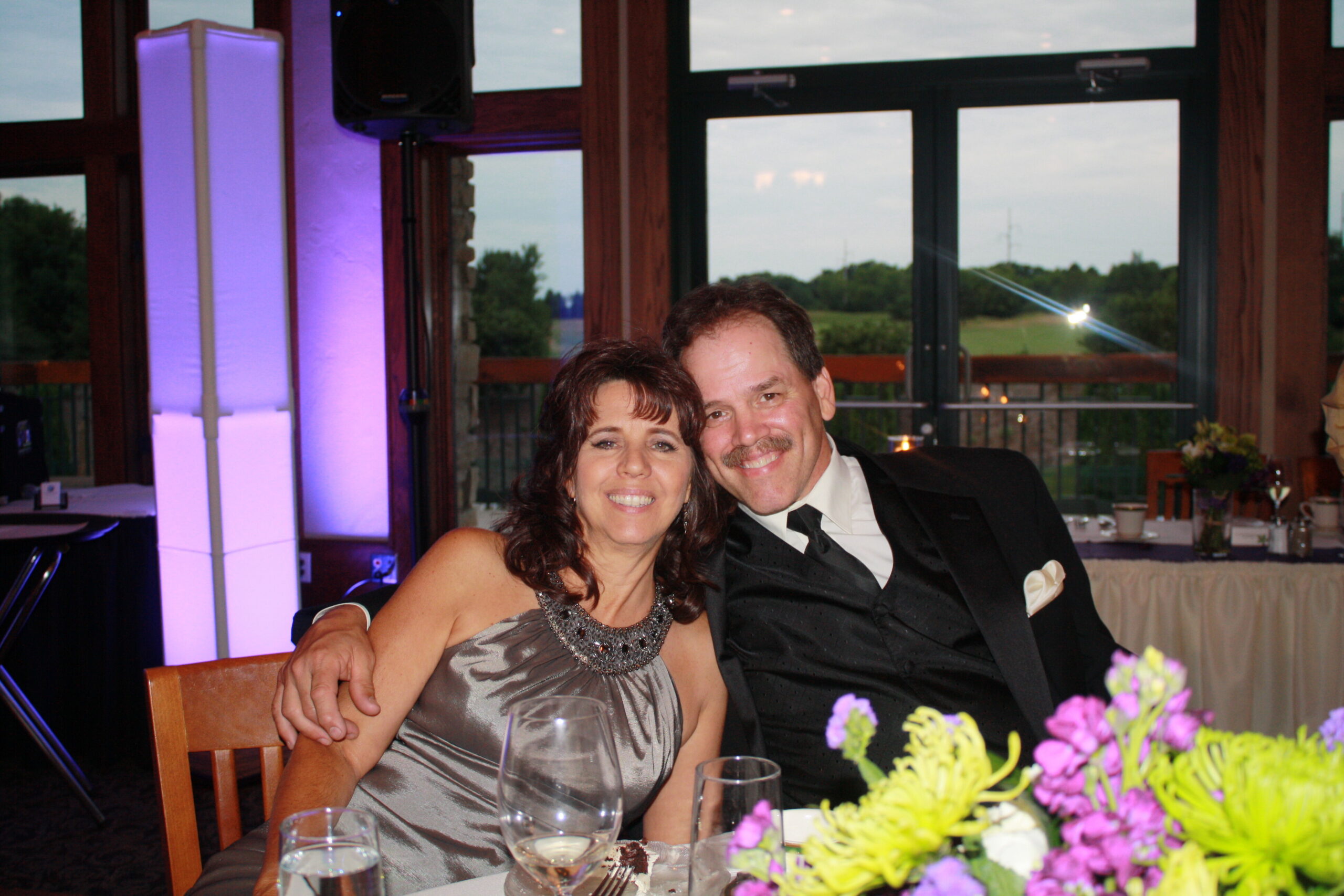 Smiling couple posing at a reception table, woman in a silver dress and necklace, man in a black tuxedo, with colorful floral centerpiece in the foreground and purple-lit columns in the background.