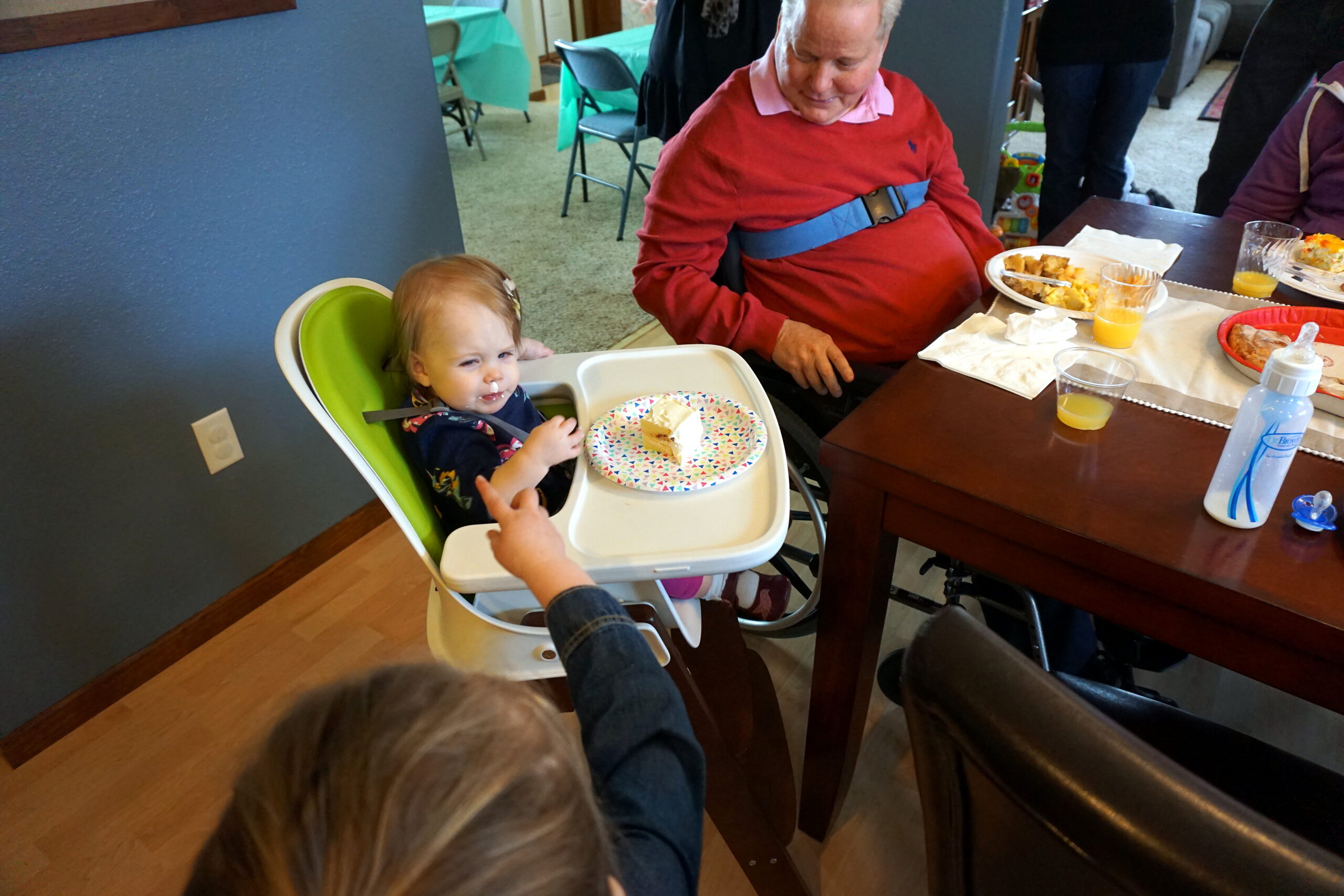 Toddler in a green high chair at a dining table with cake on a colorful plate, being reached for by adults nearby.